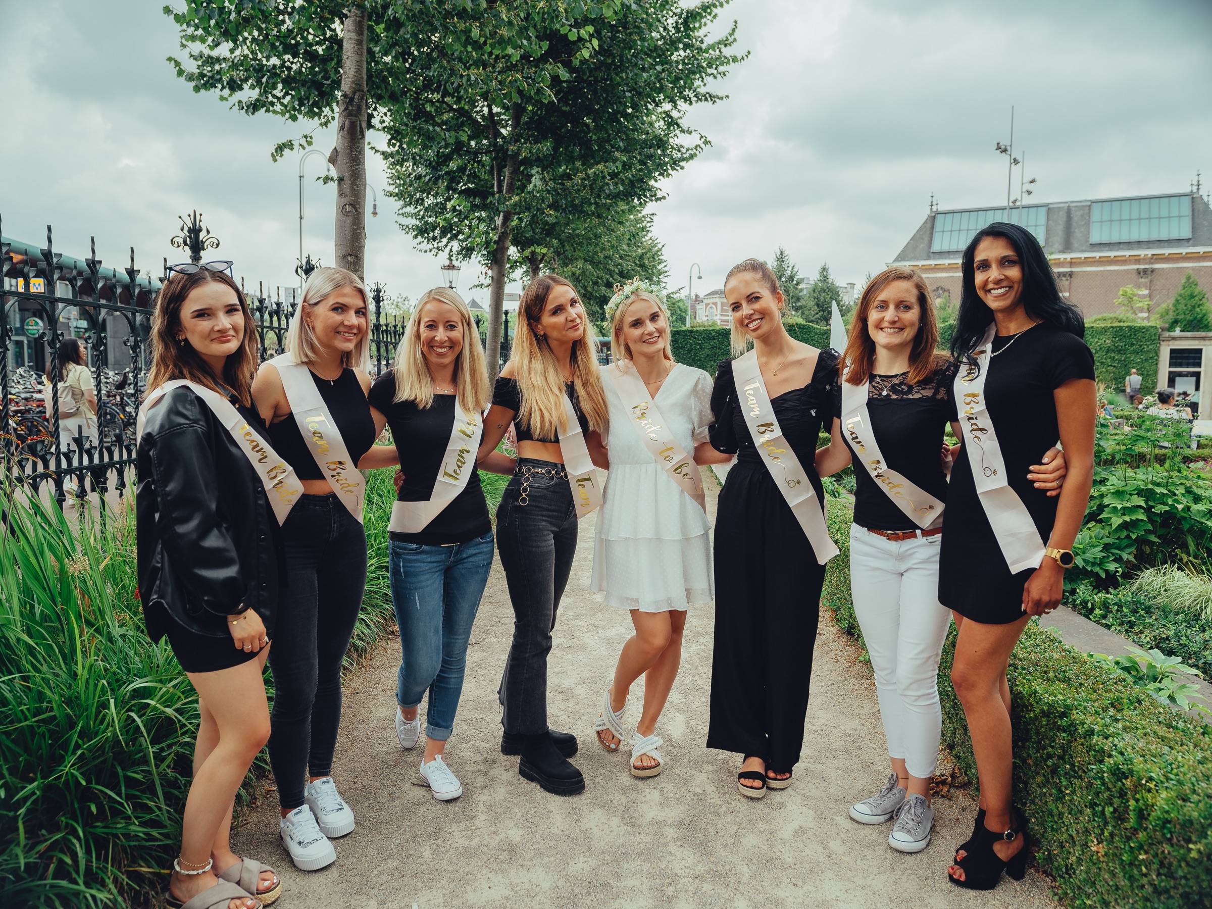 Eight women in sashes standing arm in arm on a sandy path in the Rijksmuseum Gardens, green hedges and an iron fence beside them