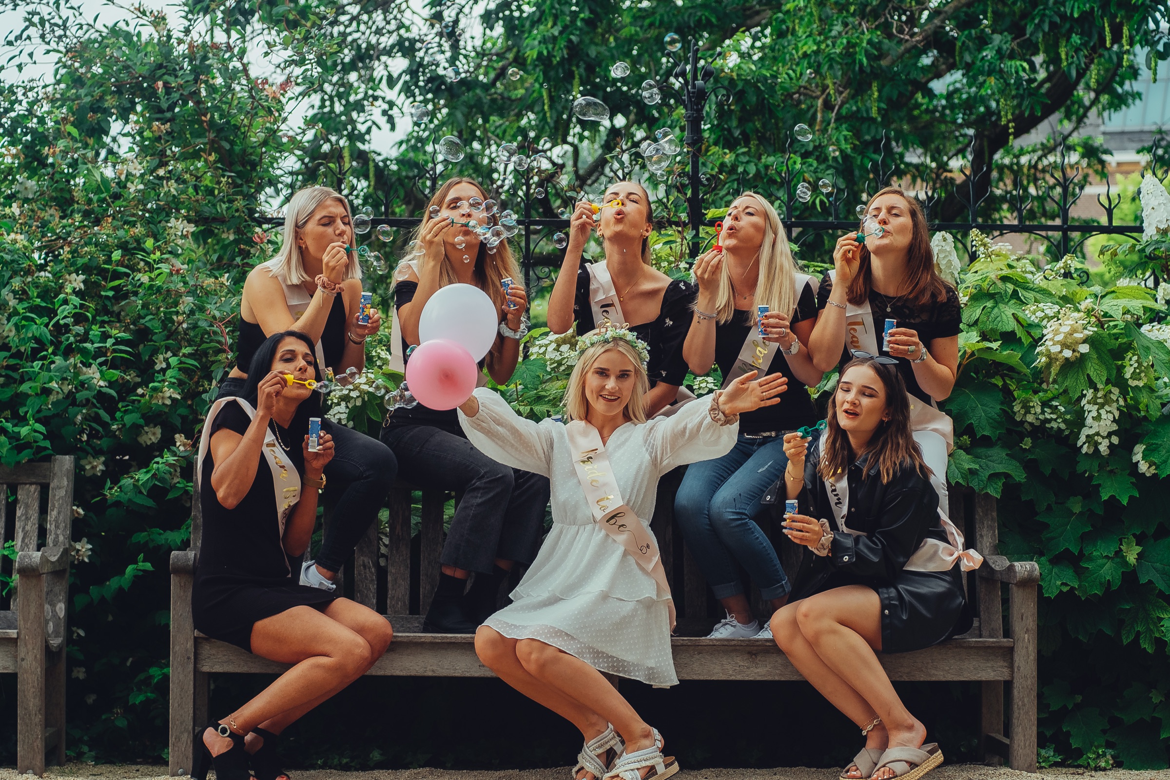 Bachelorette friends blowing soap bubbles on a bench in the Rijksmuseum Gardens, the bride in front with open arms and two balloons