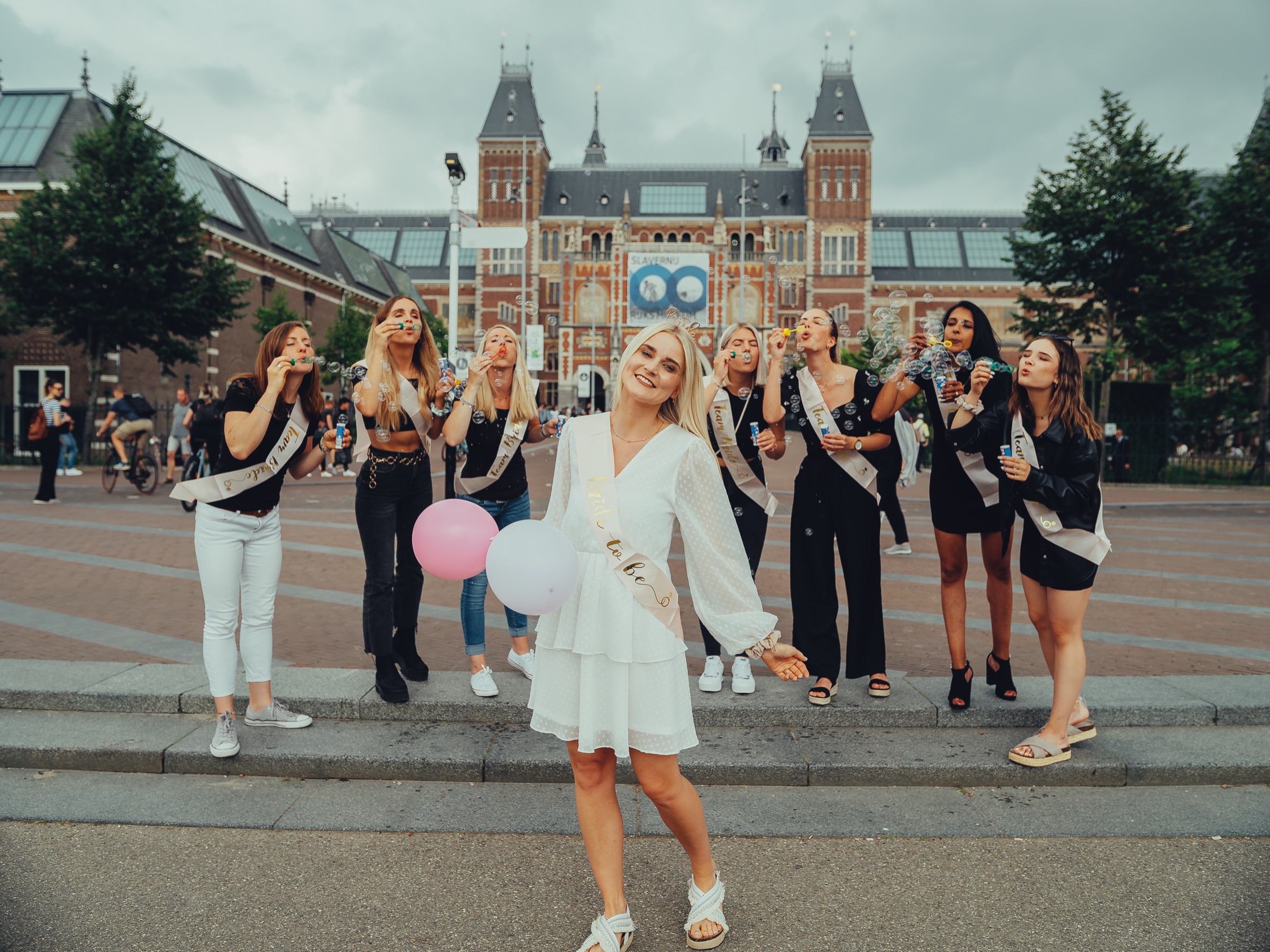 Bachelorette group blowing soap bubbles on Museumplein, the bride in a white dress and sash in front of the Rijksmuseum