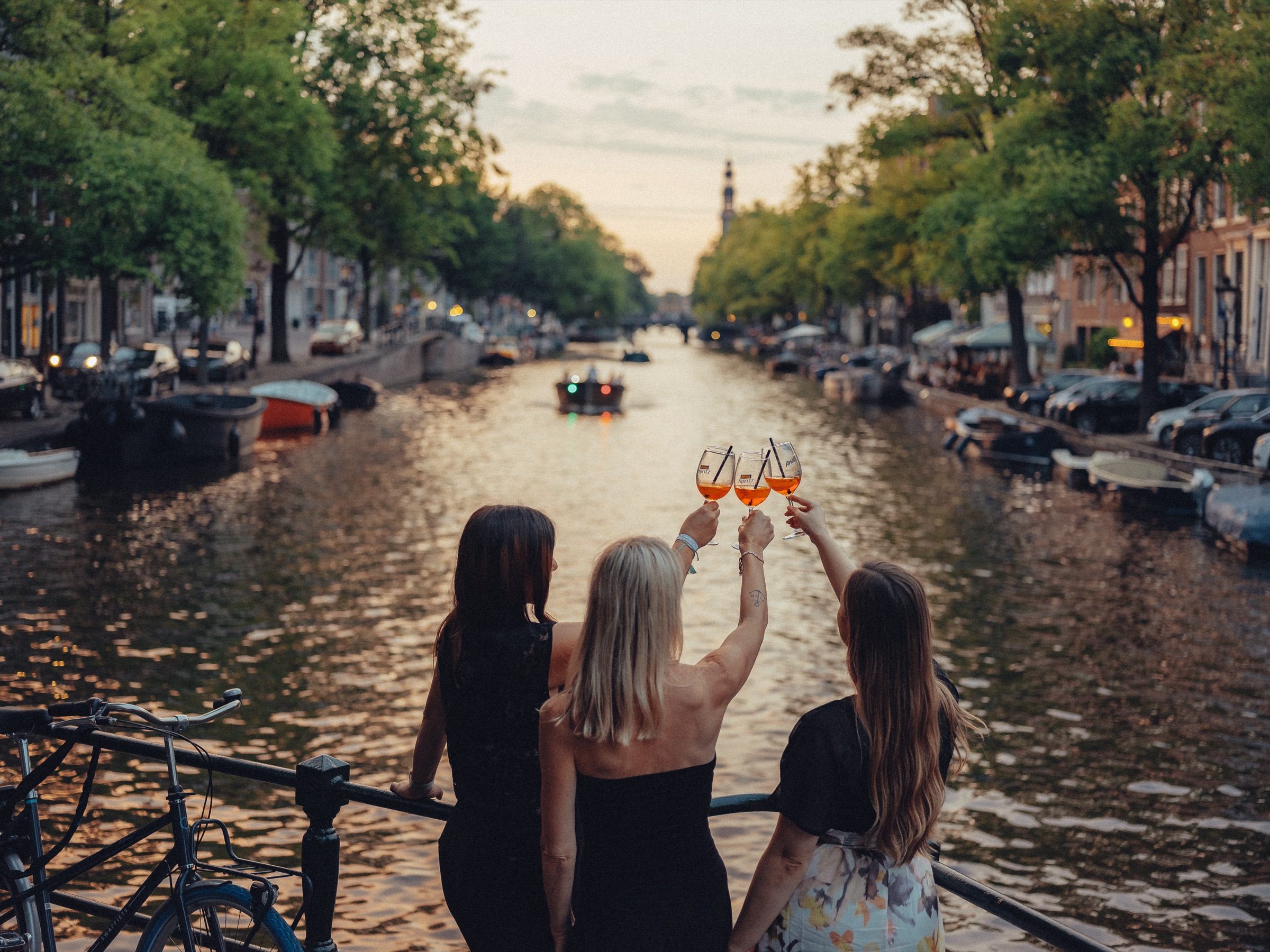 Three friends seen from behind raising spritz glasses over the canal at sunset on their bachelorette party, a church tower far away