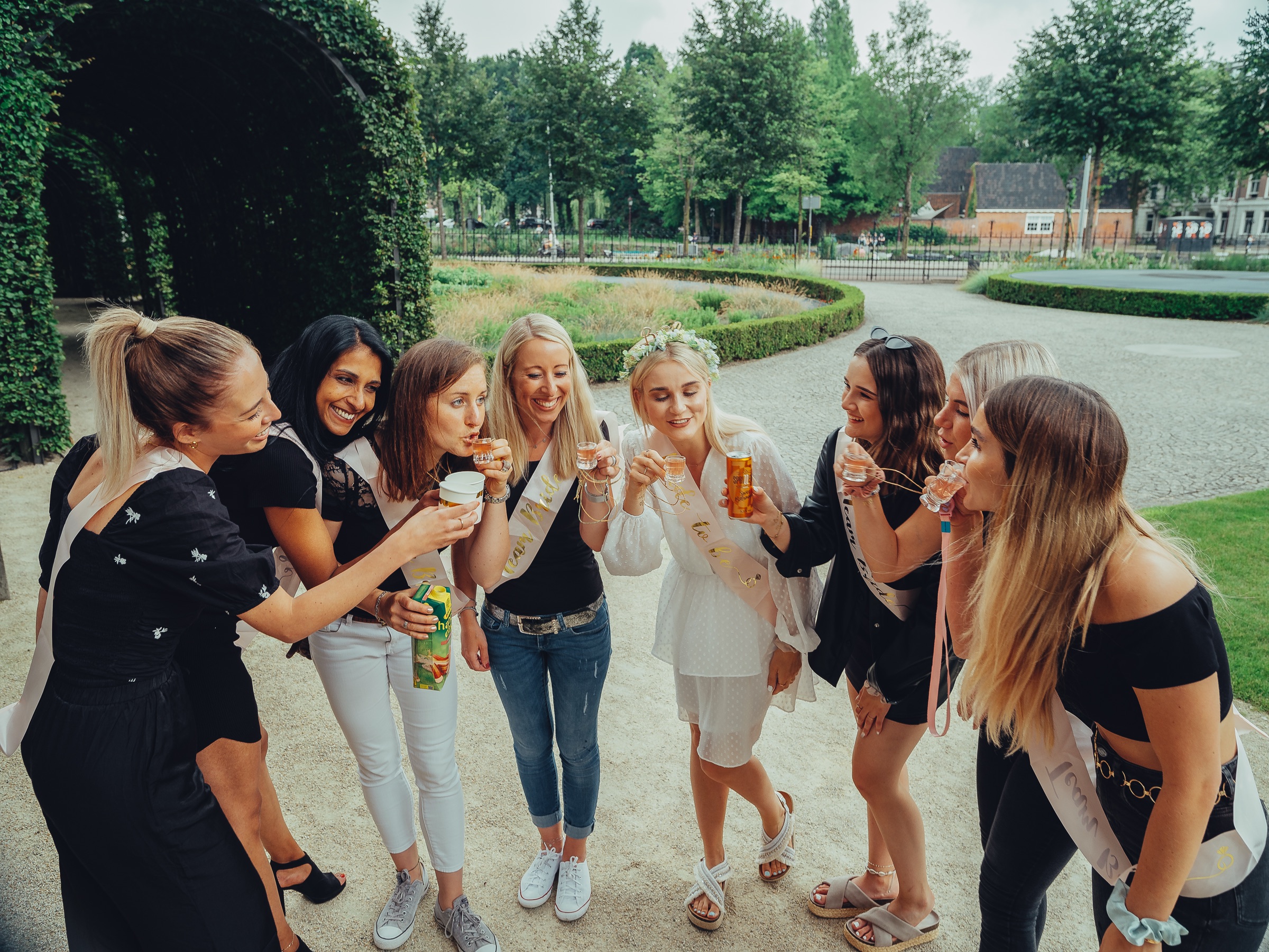 Bachelorette toast with tiny glasses on cords in the Rijksmuseum Gardens, the bride in a flower crown laughing among friends