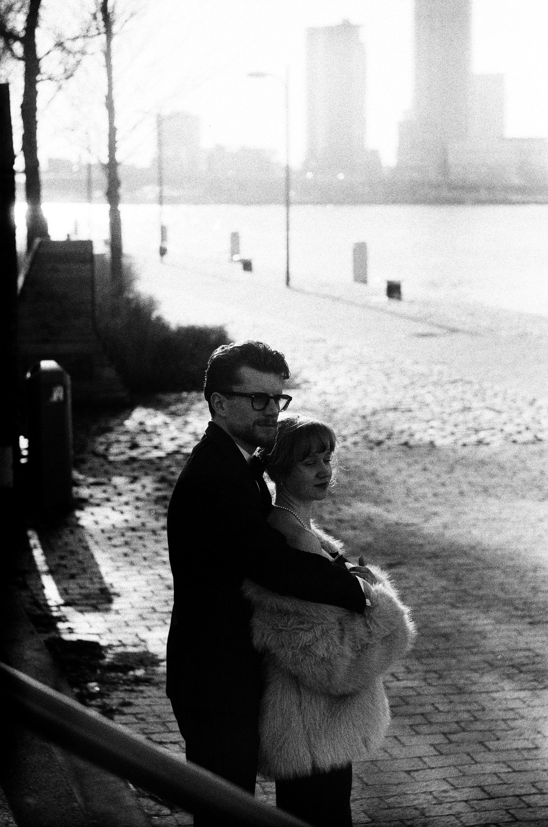 Backlit couple on a cobbled riverside quay, he in glasses and a tuxedo, she in a pale fur coat, the river and hazy towers behind