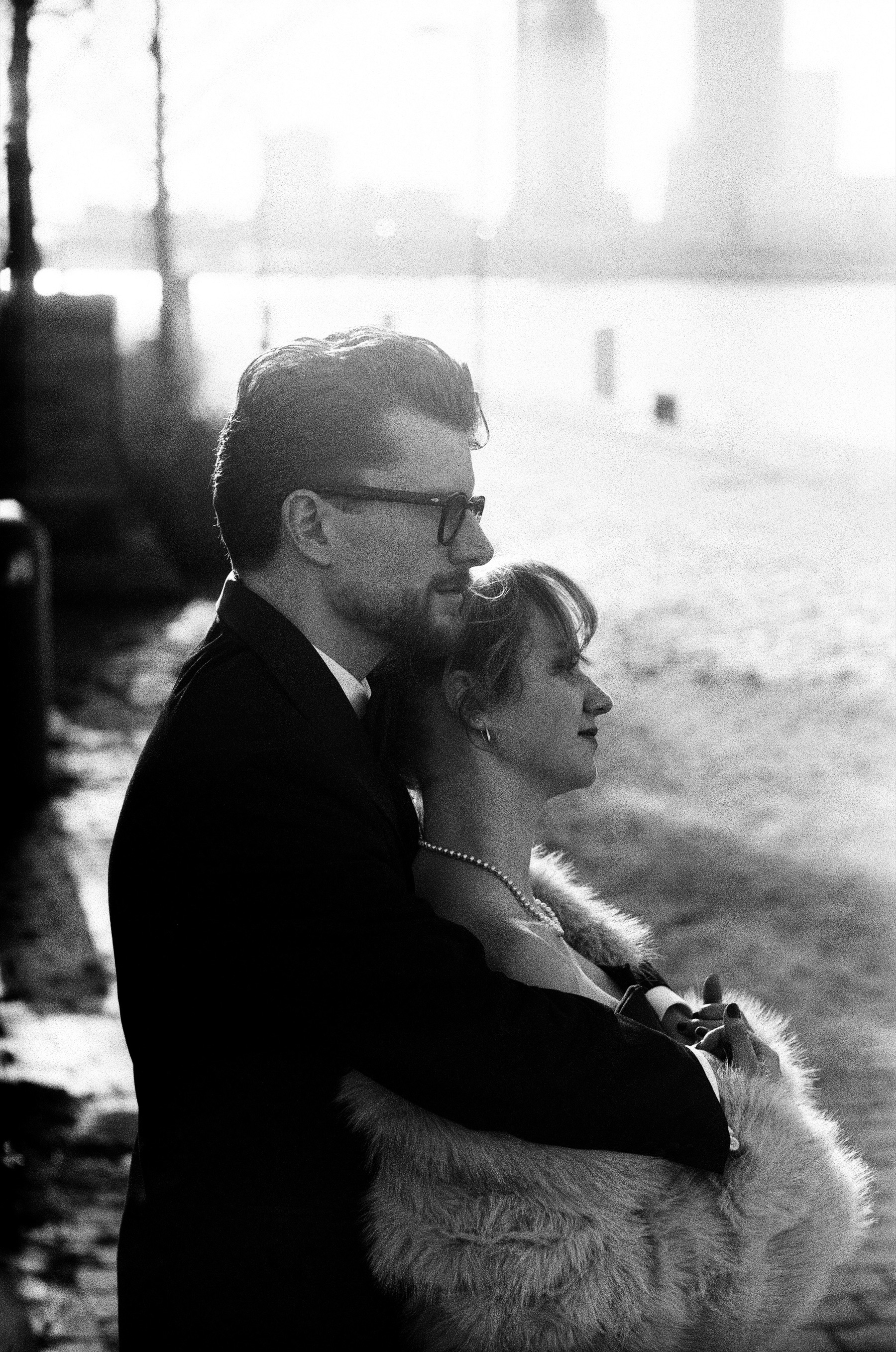 Man in a dark suit holding his partner from behind by the Maas in Rotterdam, the river shining in backlight and towers in a haze, on Ilford Delta 100