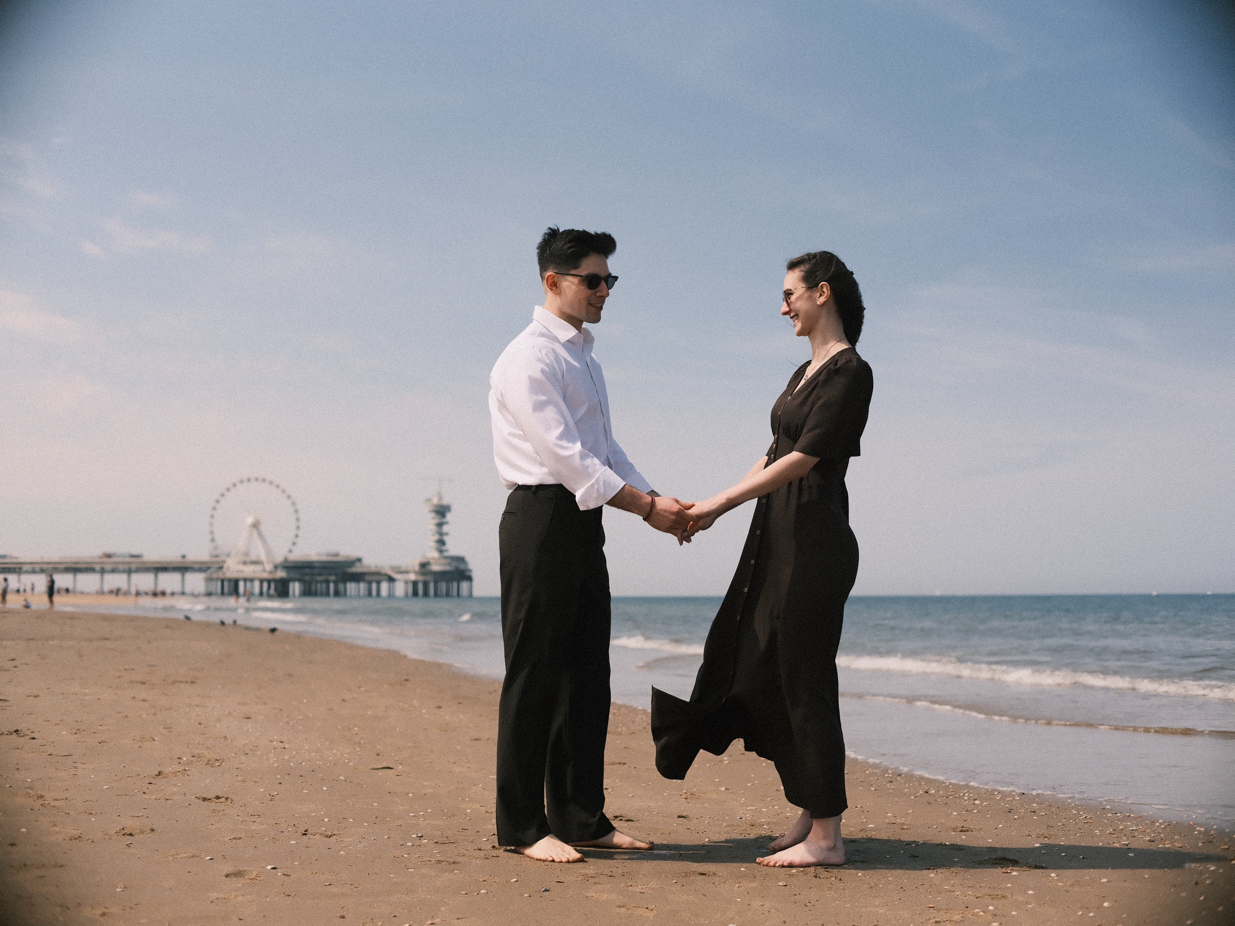 Barefoot couple holding hands on Scheveningen beach before the proposal, the Pier and its Ferris wheel on the horizon