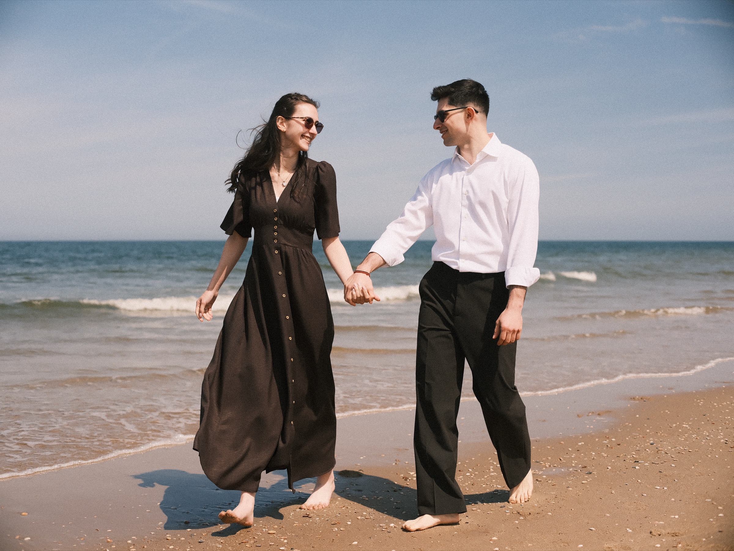 Barefoot couple walking hand in hand at the water edge on Scheveningen beach before the surprise proposal, her long dark dress swinging