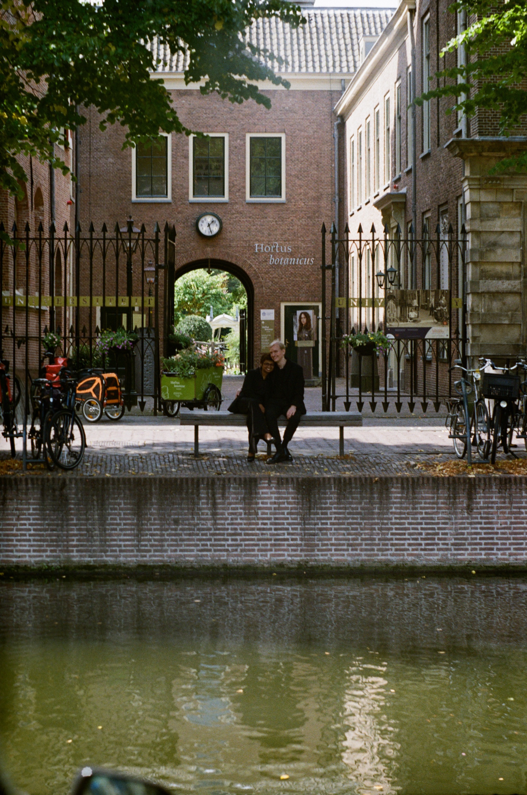 Couple on a bench before the Hortus botanicus gate across the Rapenburg in Leiden, green canal water in the foreground