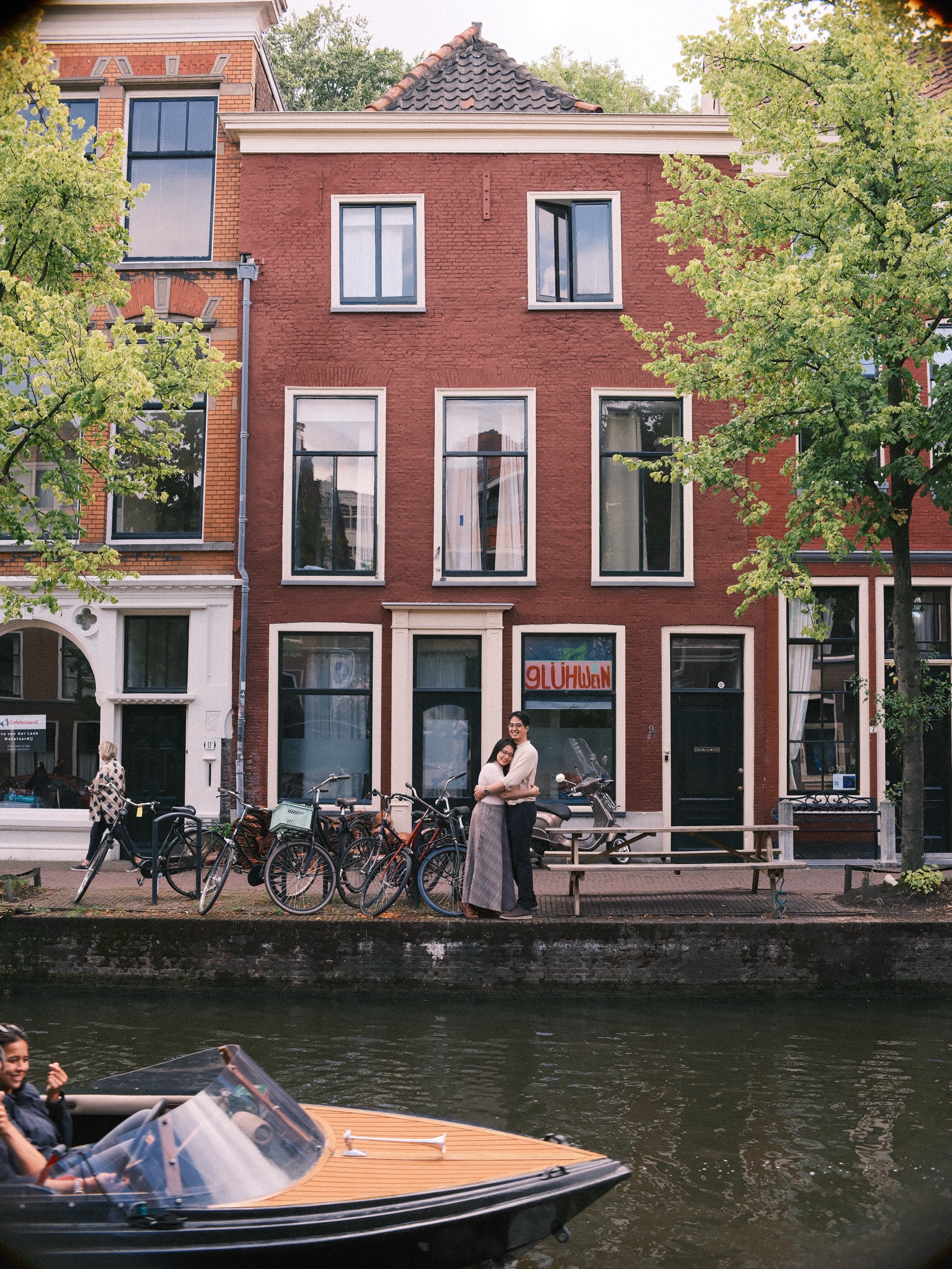 Couple on a canal quay in front of a red brick house while a small wooden boat with a windshield passes in the foreground