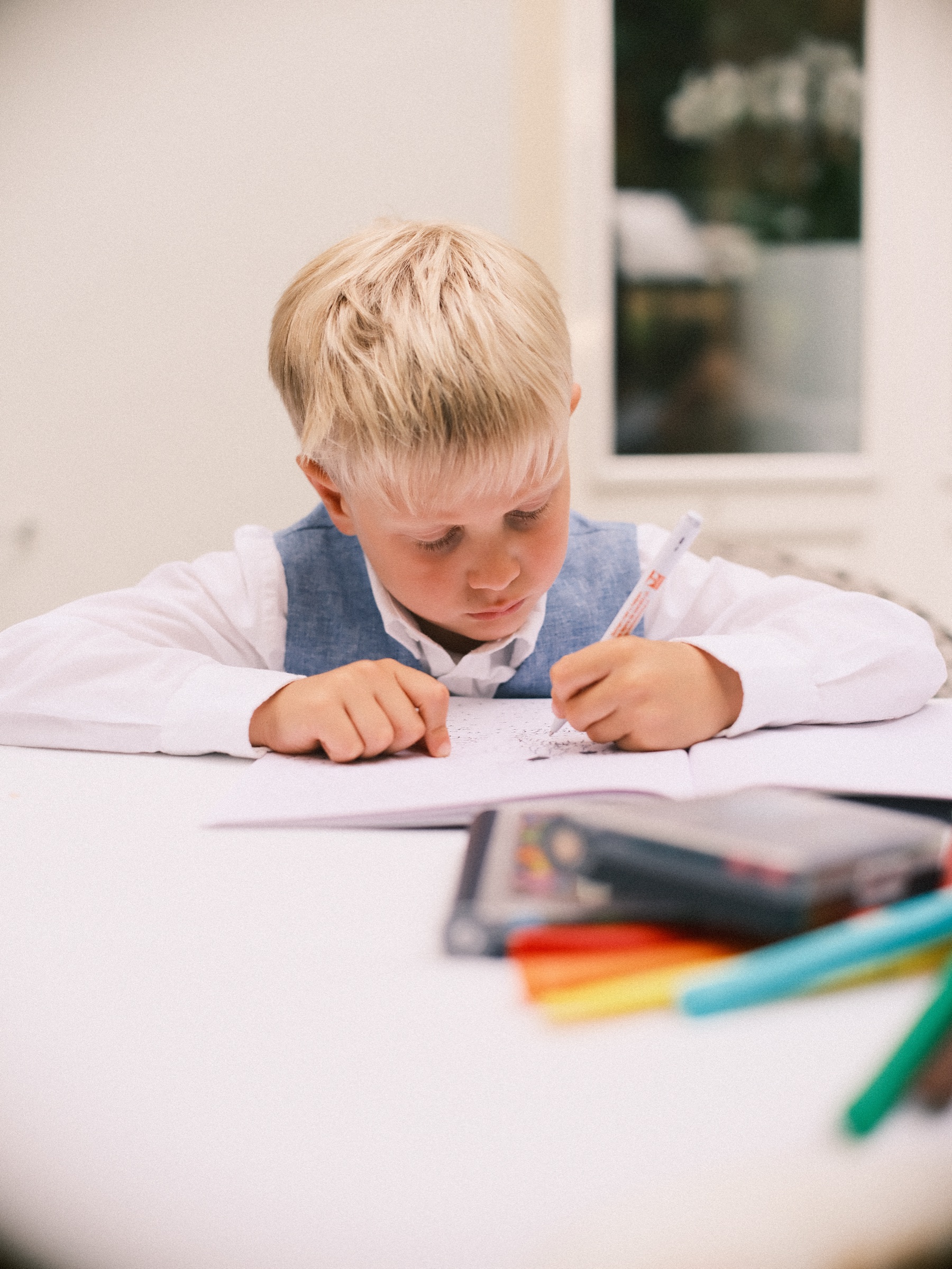 Jongen in een wit overhemd en blauw gilet tekent met gekleurde stiften aan een witte tafel tijdens het trouwfeest
