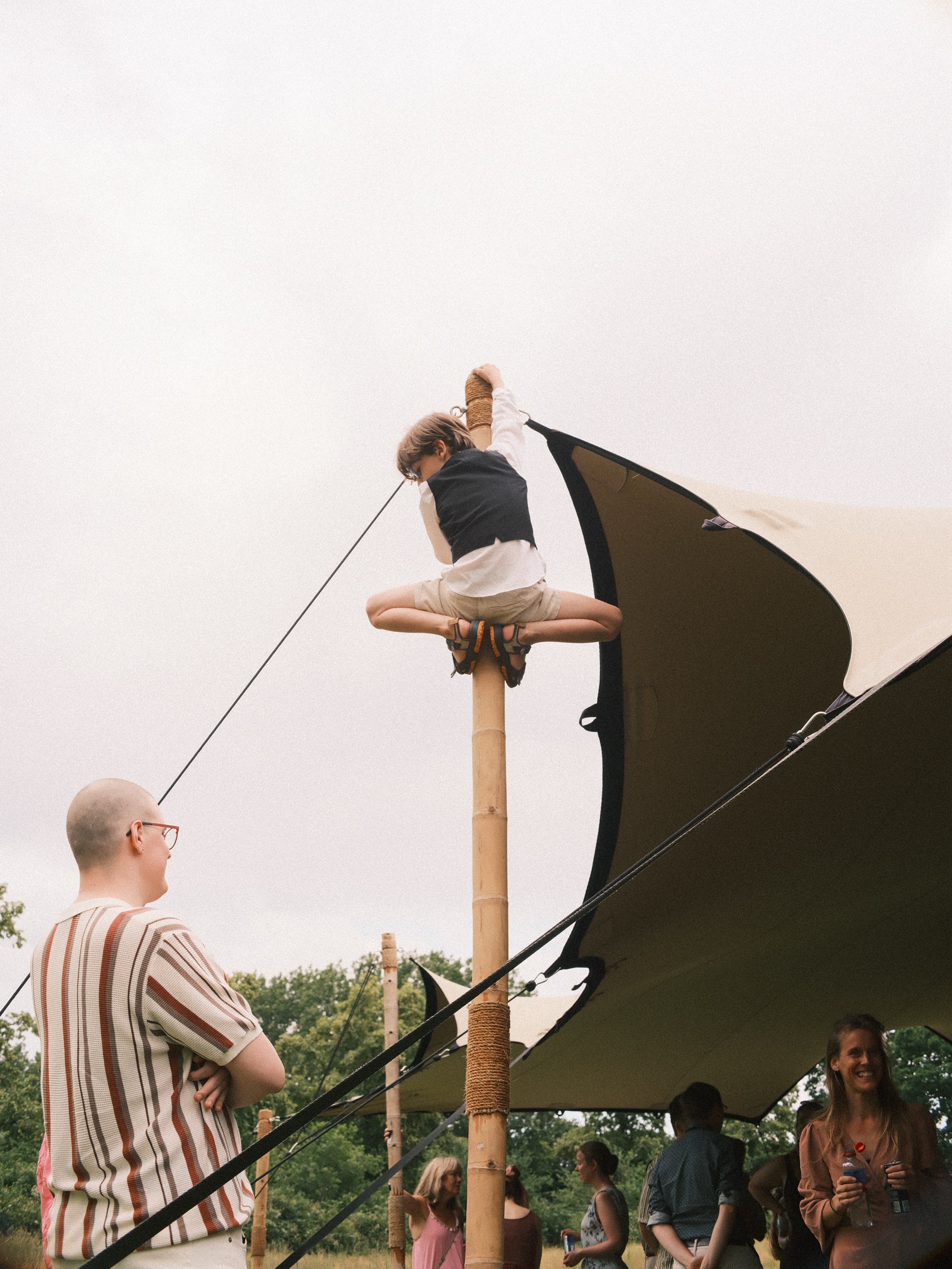 Boy perched at the top of a tall wooden tent pole beside the canvas, a guest in a striped shirt watching with folded arms