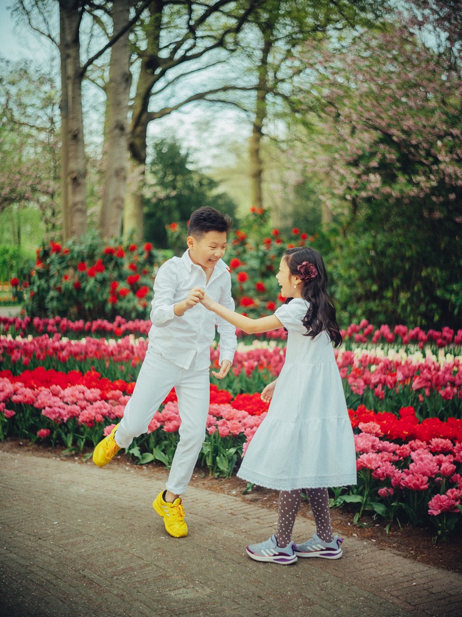 Jongen met gele sneakers laat zijn zusje in een witte jurk en stippenpanty rondtollen op een pad tussen rode en roze tulpen