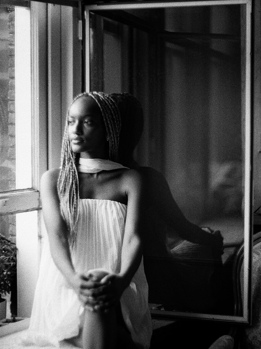 Window light falls on a woman with long braids and clasped hands in this black-and-white film portrait