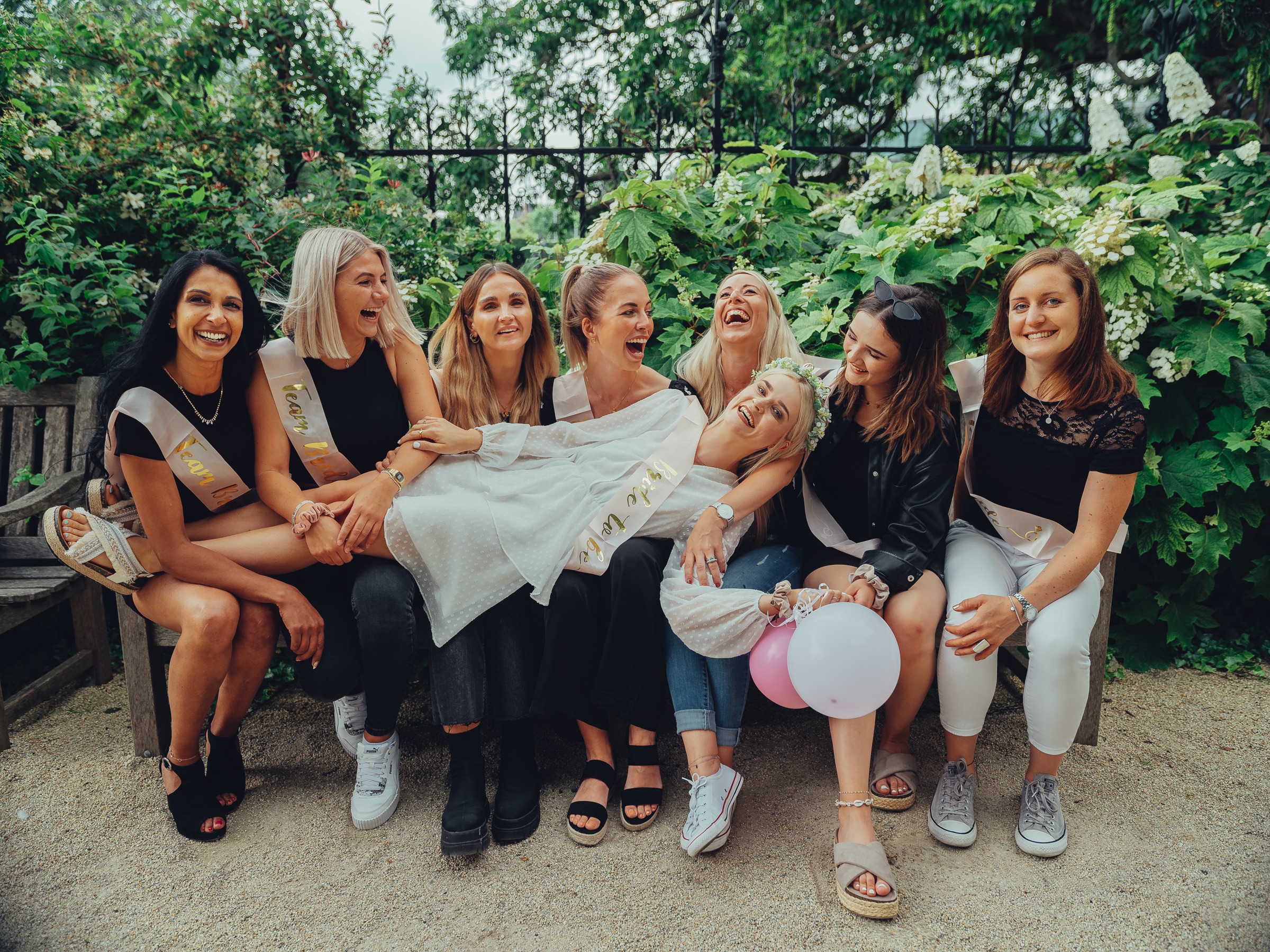 Bride laughing as she lies across her friends' laps on a wooden bench in the Rijksmuseum Gardens, white flowers behind