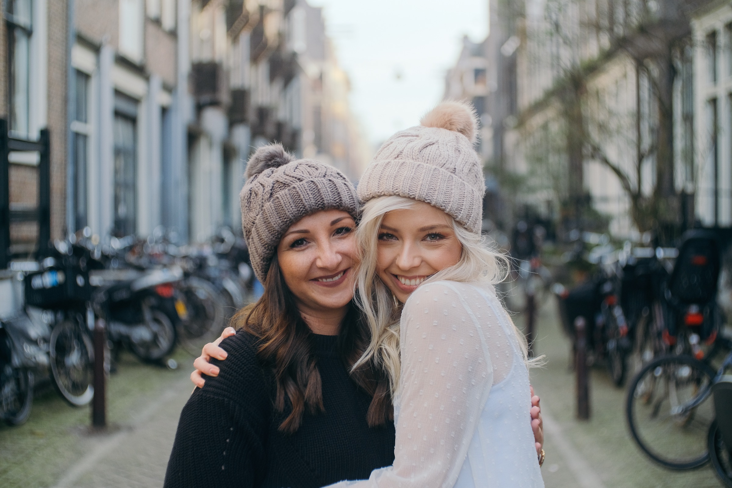 Bride and a friend hugging cheek to cheek in cable-knit beanies with pompoms, both smiling straight into the camera