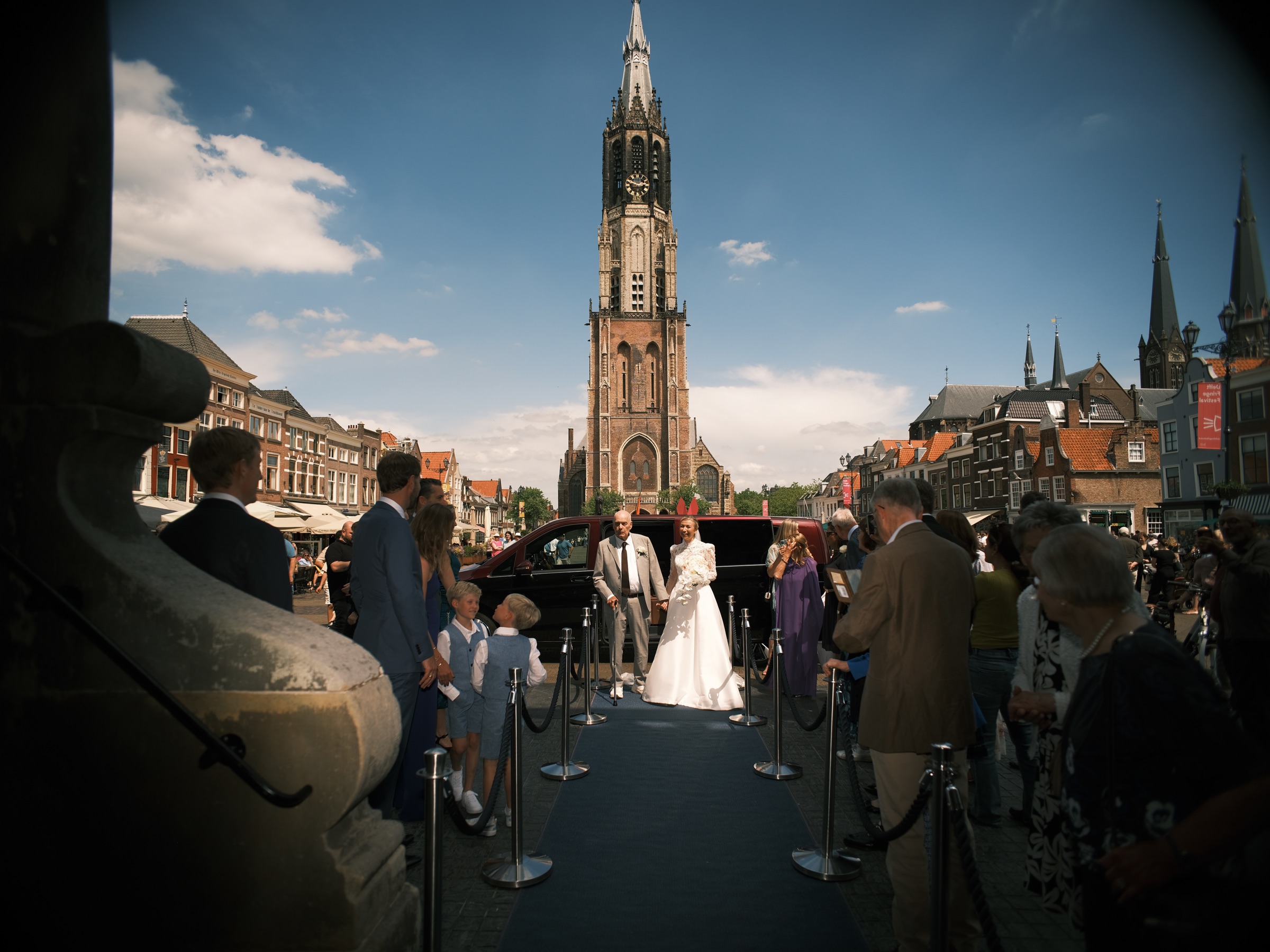 Bruid op de blauwe loper voor het stadhuis, met gasten aan weerszijden en recht vooruit de toren van de Nieuwe Kerk in Delft