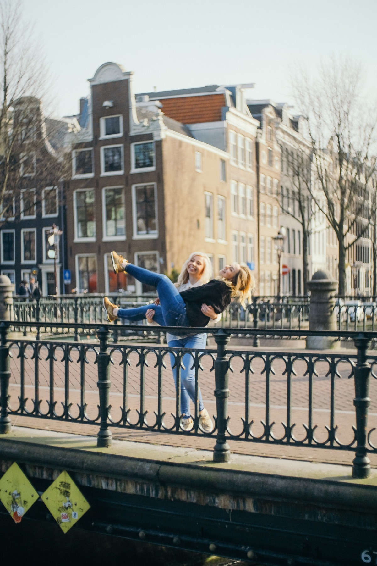Bride carrying a laughing friend in her arms on a canal bridge, gold sneakers kicking up behind the iron railing