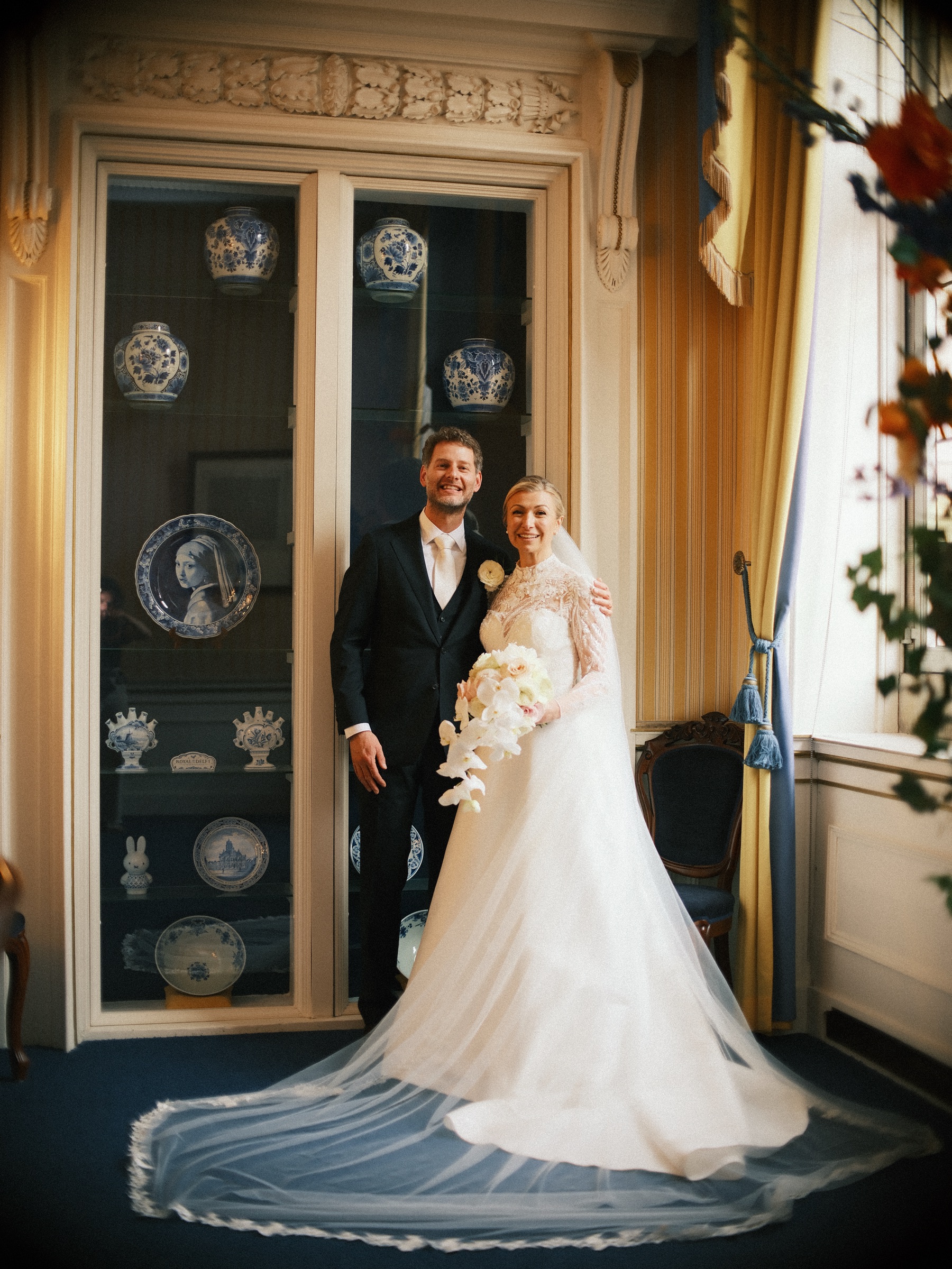 Bruid en bruidegom bij een vitrinekast vol Delfts blauw in het stadhuis van Delft, haar lange sluier uitgespreid over de blauwe vloer