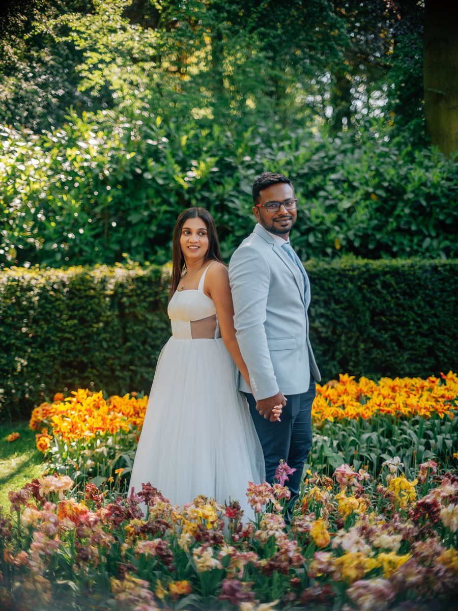 Bruid in een witte jurk met doorschijnende stukjes bij de taille staat hand in hand met de bruidegom bij een haag in Keukenhof
