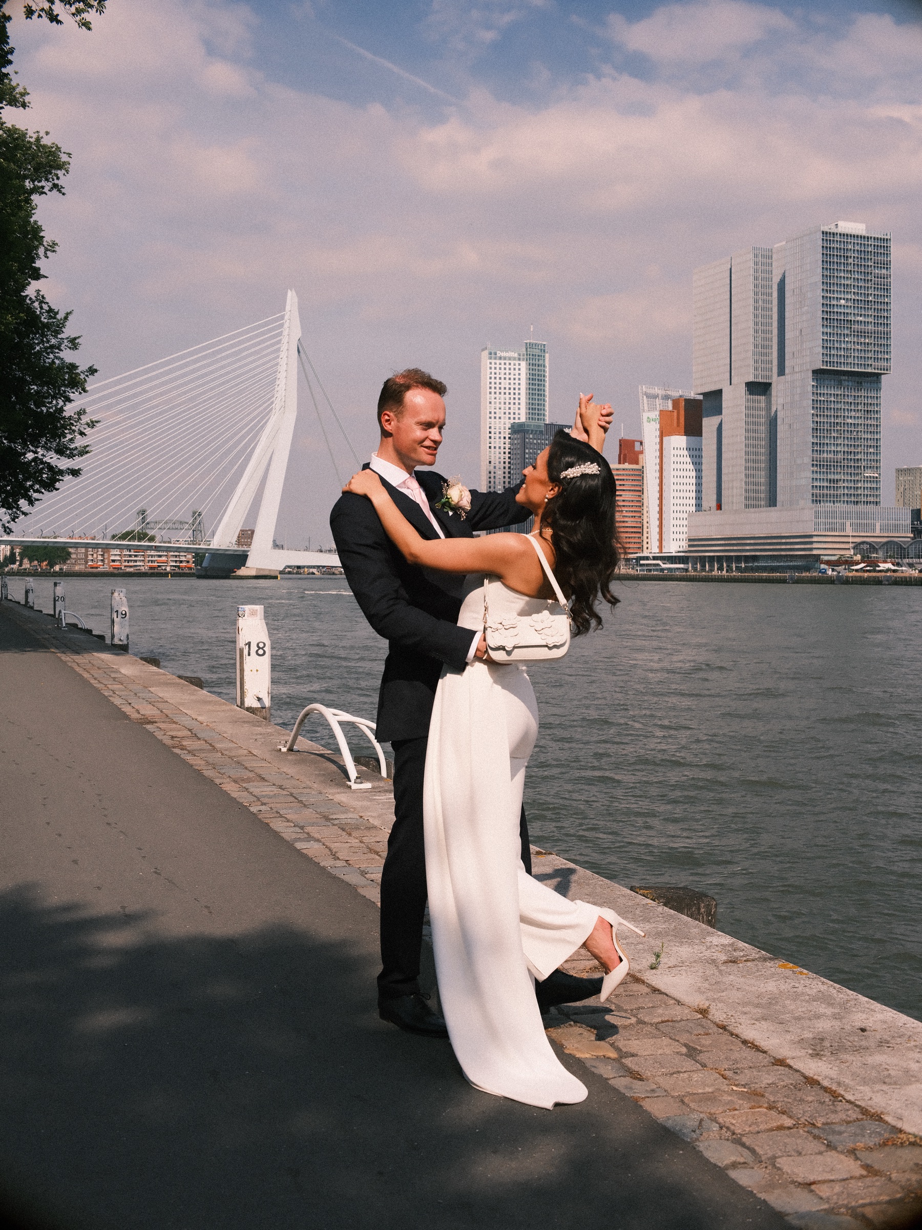 Bruid wipt een hak omhoog terwijl de bruidegom haar vasthoudt op de Maaskade, met de Erasmusbrug en hoge torens