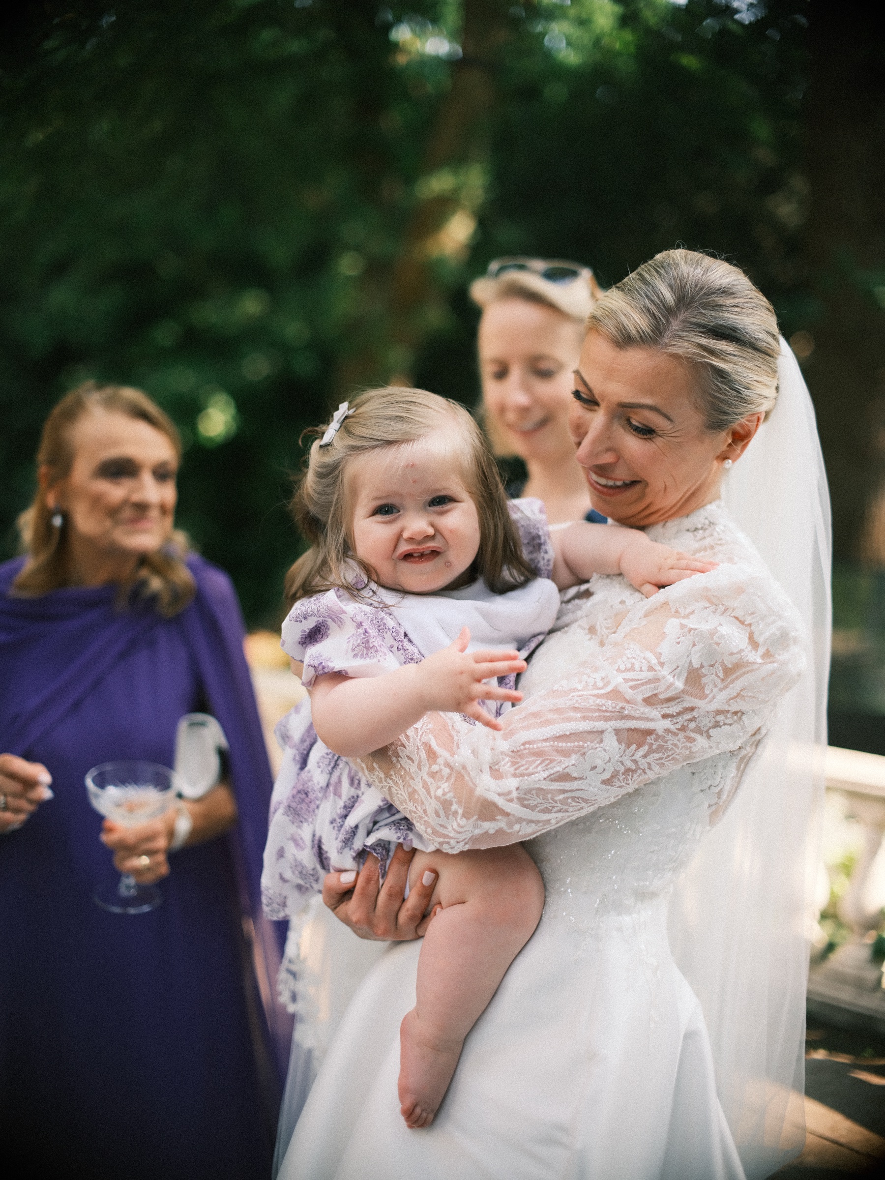 Bruid met een schaterend meisje in een jurkje met lila bloemen op het tuinfeest, met een gast in een paarse jurk ernaast