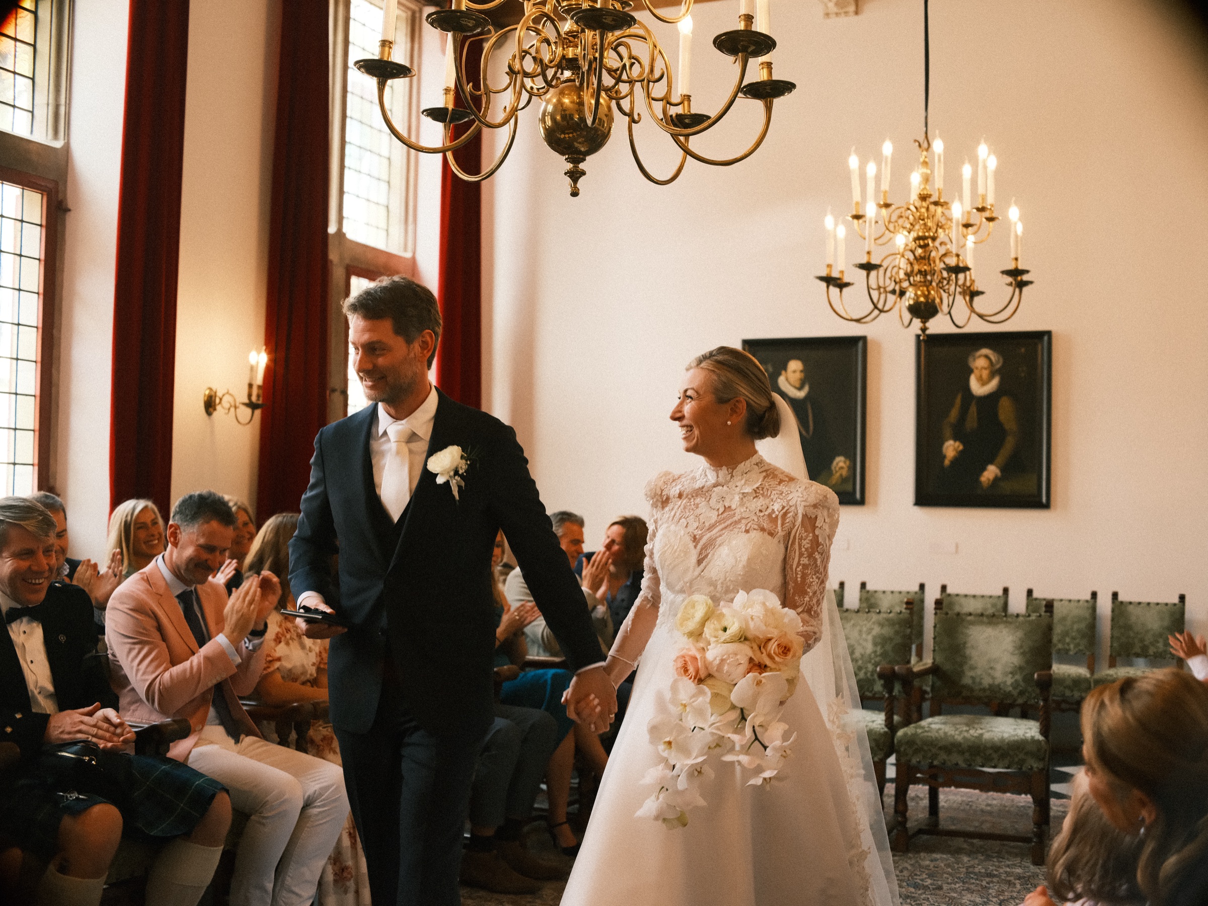 Lachende bruid loopt met haar boeket van witte orchideeën terug door het gangpad, met de bruidegom voor haar en klappende gasten