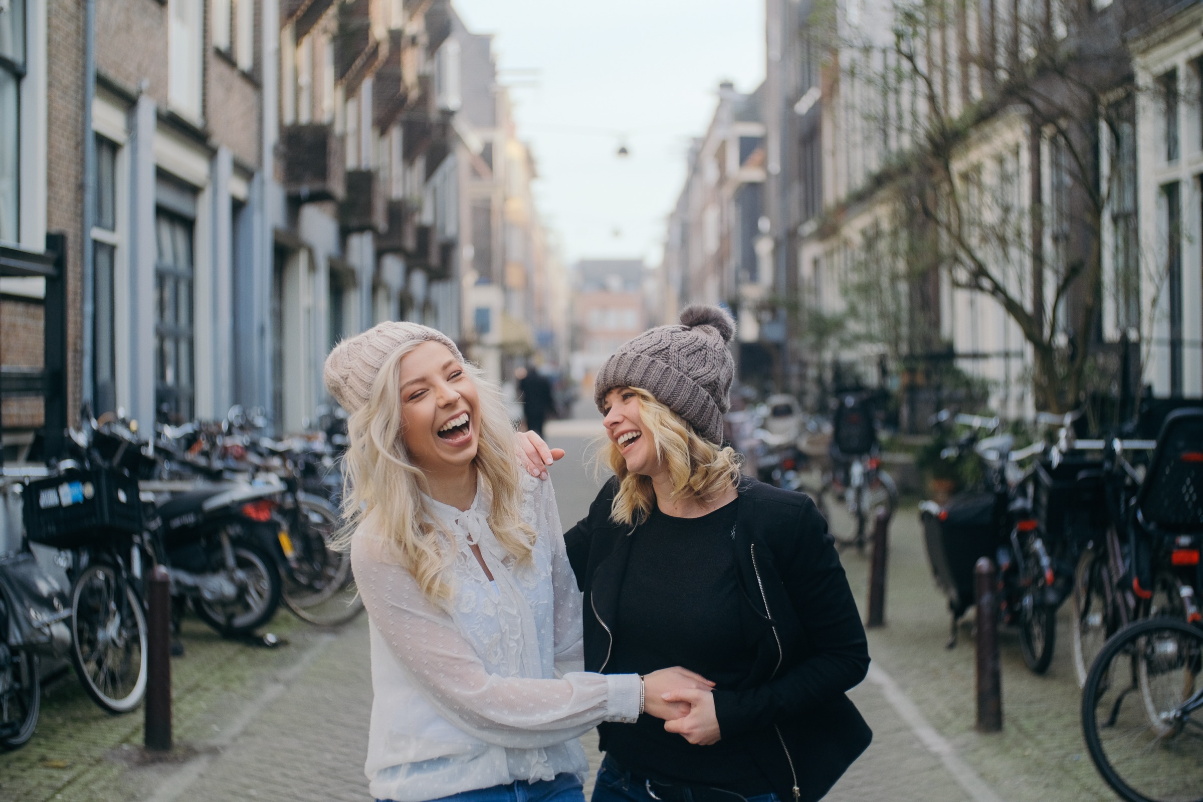 Bride laughing hand in hand with a friend in a black jacket, bikes parked along the narrow street behind them