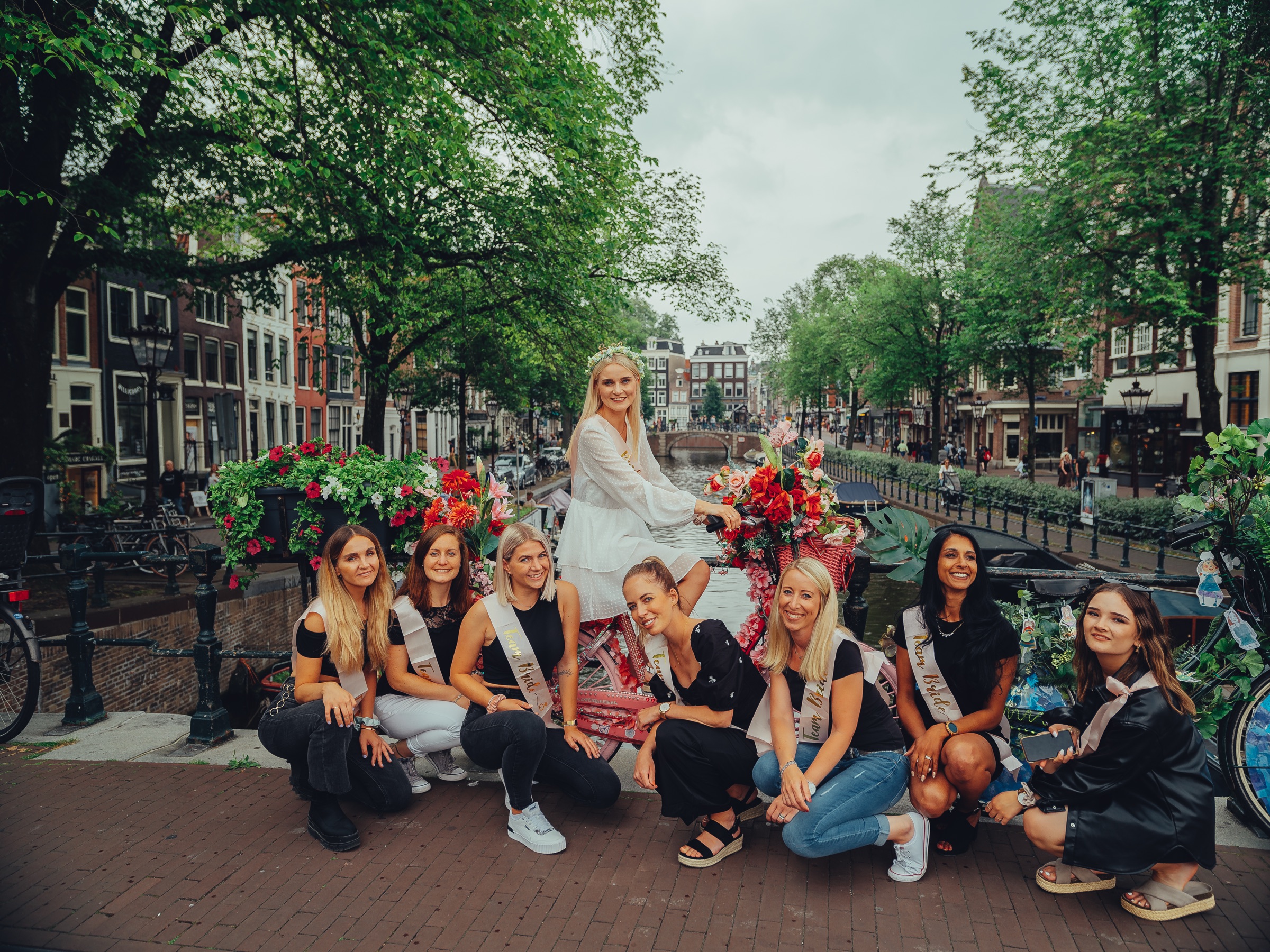 Bride-to-be sitting on a flower bike above her crouching friends on a canal bridge, a long tree-lined canal behind