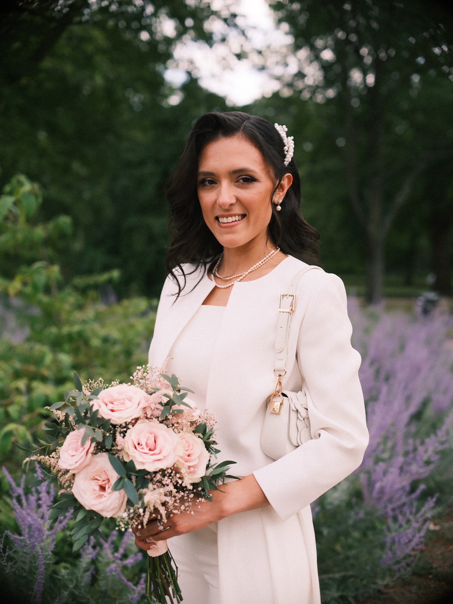 Bruid in een wit jasje met een parelketting en een parelkam houdt roze rozen vast voor paarse bloemen in Het Park