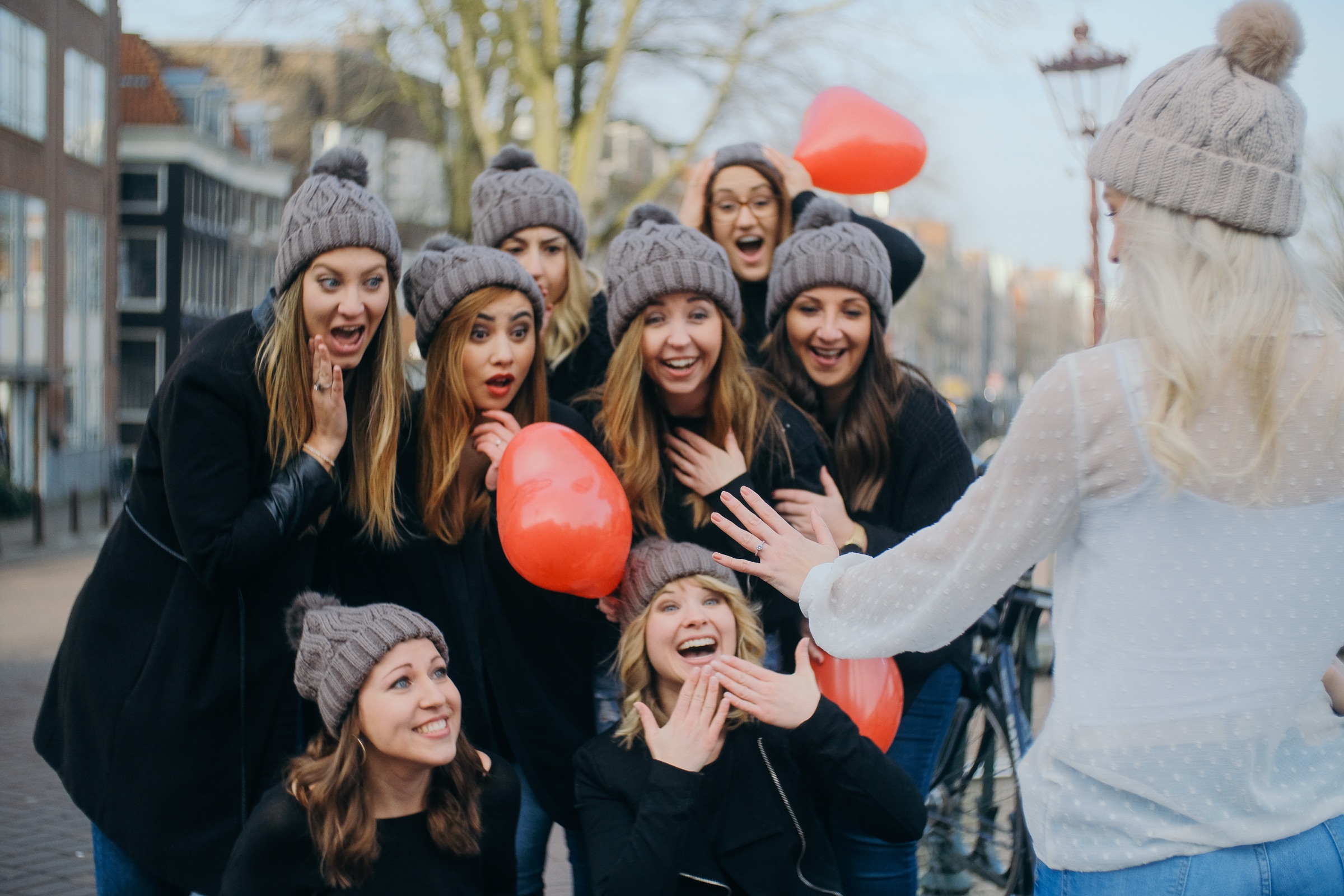 Bride holding out her hand to show her ring as friends in gray beanies gasp, red heart balloons in their hands