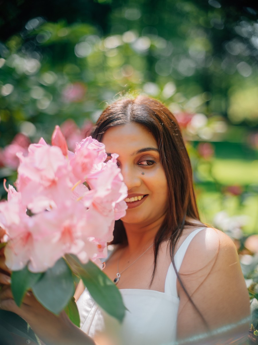 Bruid lacht vanachter een tros roze rododendronbloemen, met haar lange donkere haar over haar witte jurk