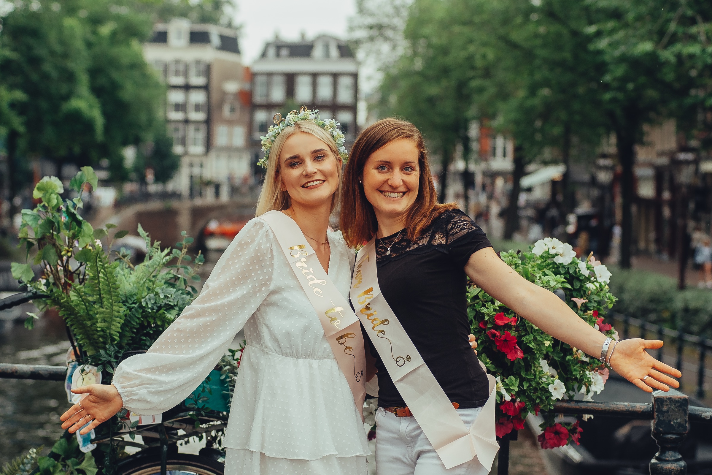 Bride-to-be in her sash and a friend in a Team Bride sash spreading their arms wide on a flower-lined canal bridge
