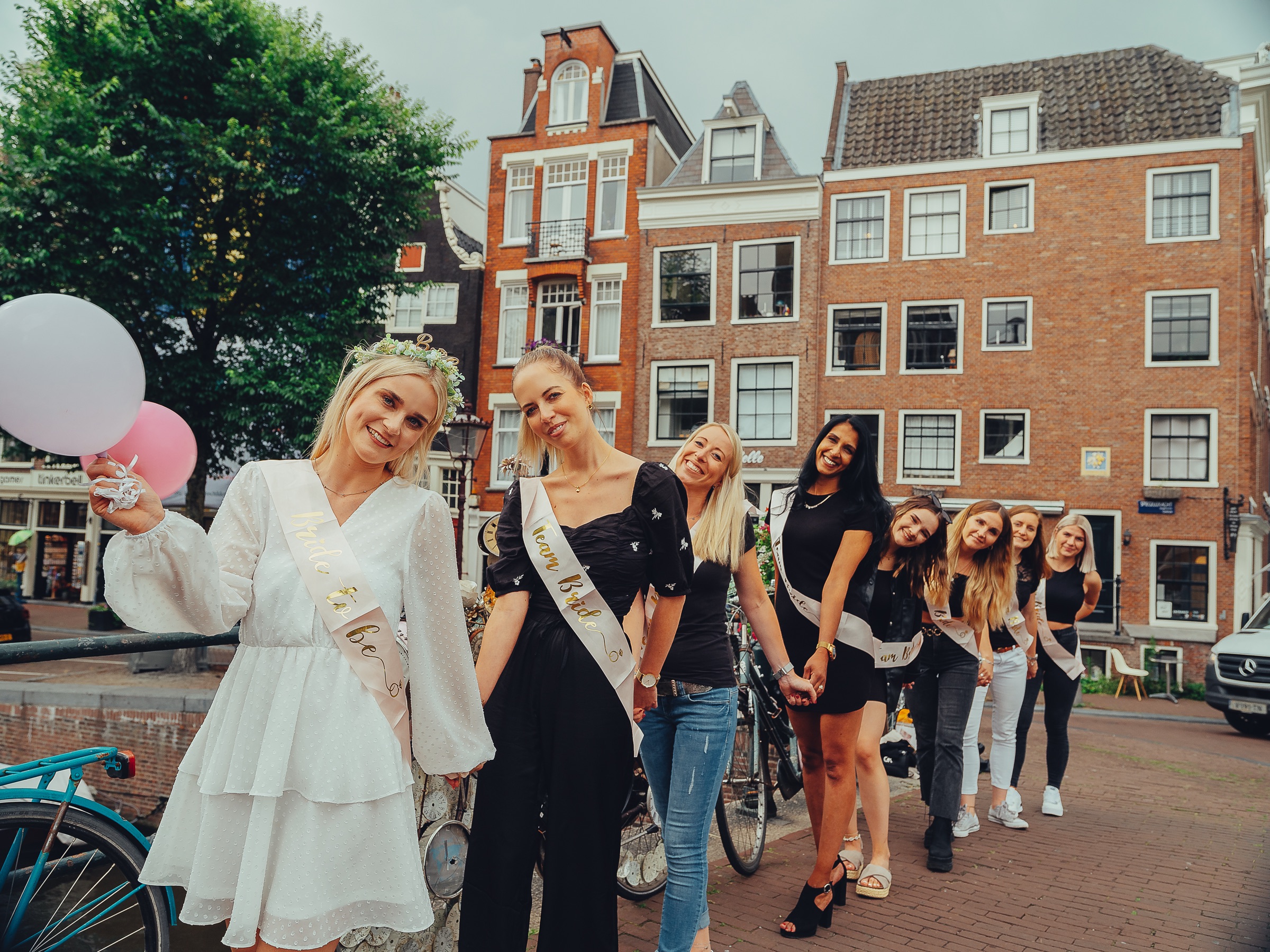 Bride-to-be with pink and white balloons leading a chain of friends holding hands along a canal, brick houses behind