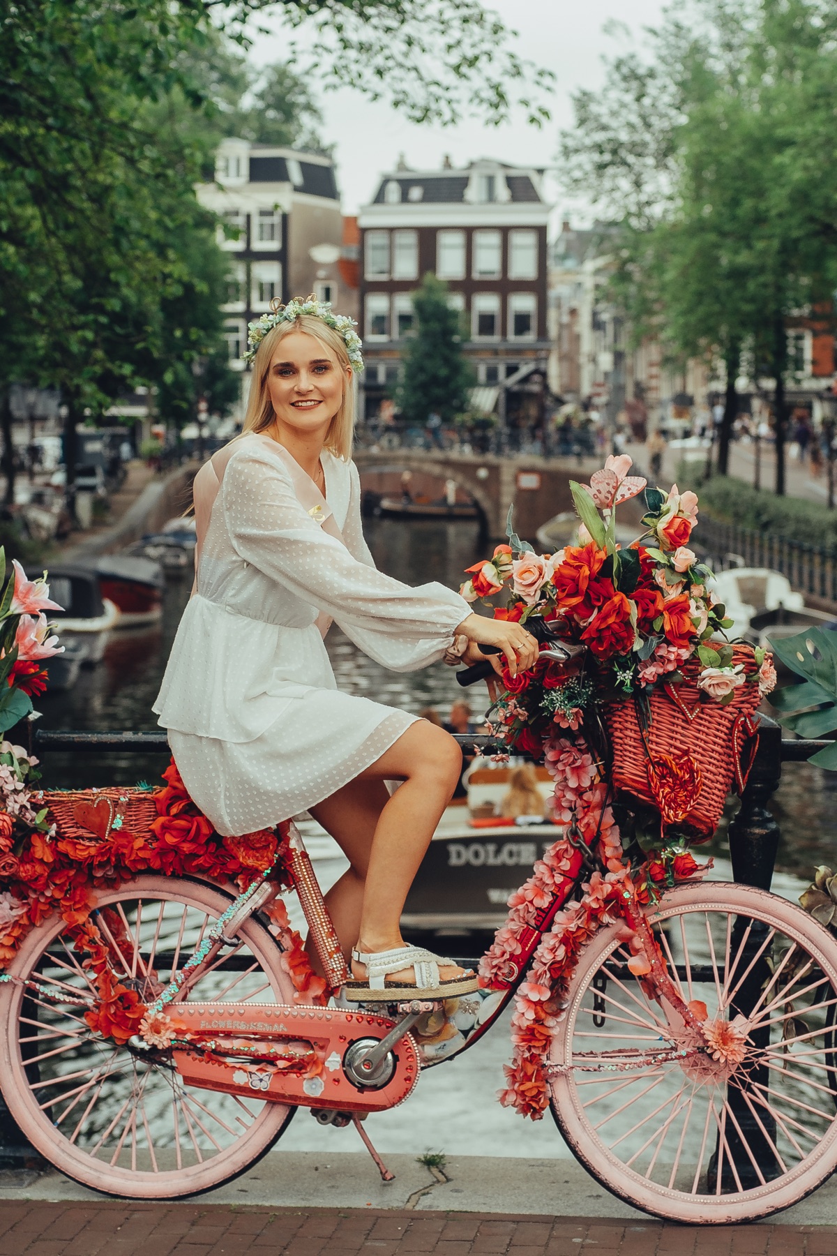 Bride-to-be in a white dress and flower crown on a bike covered in red and pink flowers, on a canal bridge with boats behind