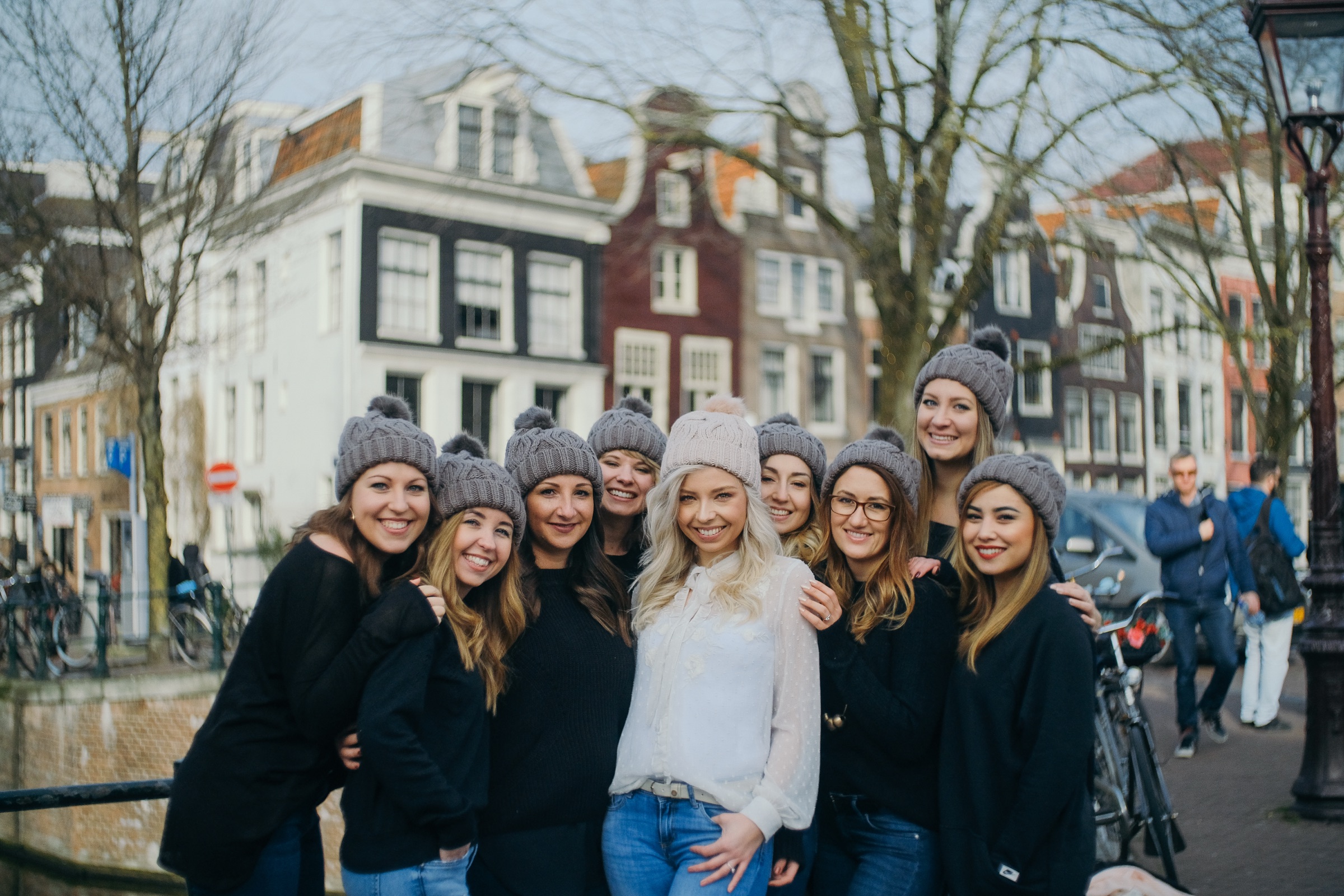 Bride-to-be in a white lace blouse and cream beanie hugged by friends in gray beanies beside an Amsterdam canal, bare trees behind