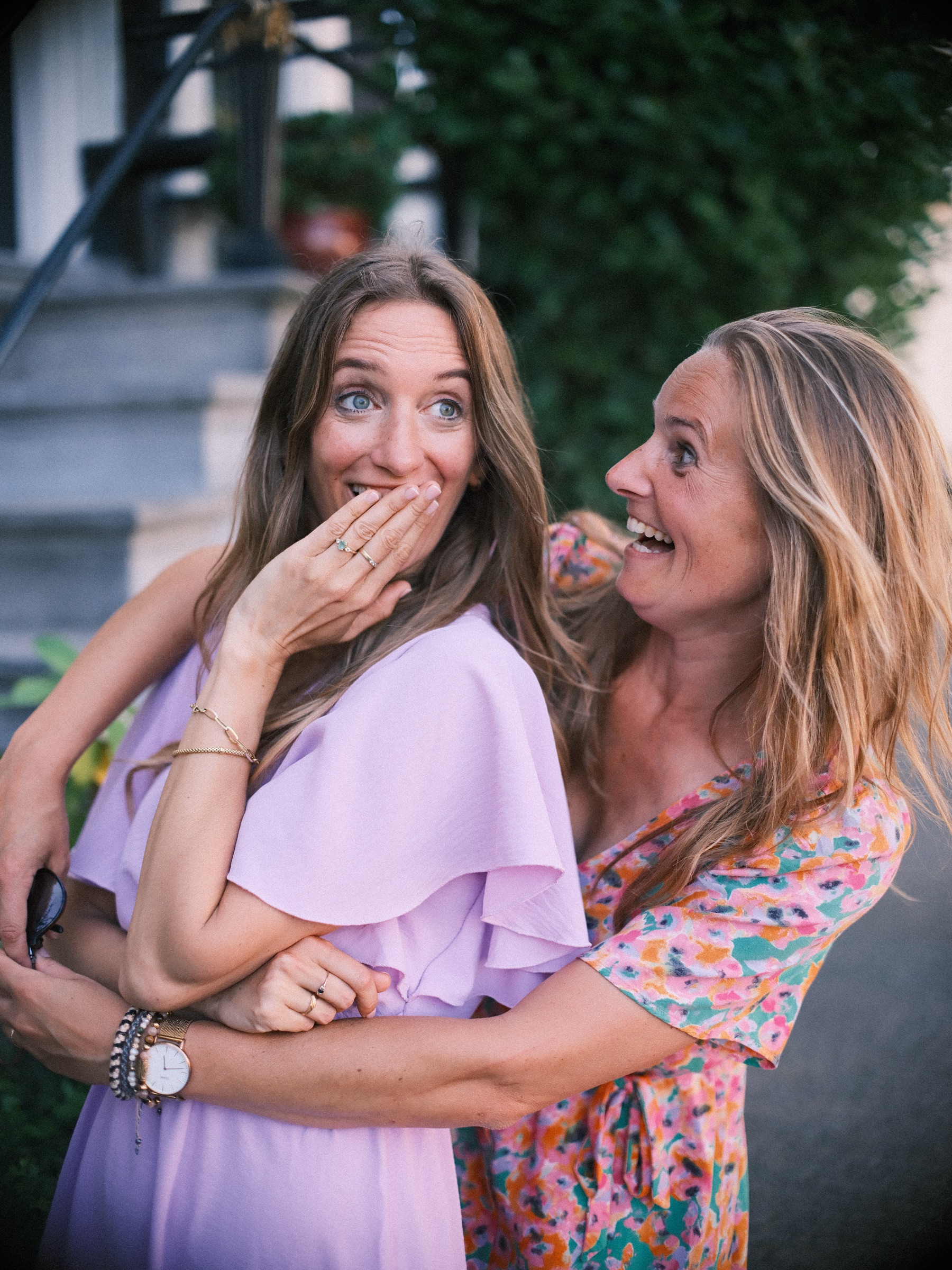 Bride-to-be covering her mouth with laughter as a woman in a floral dress hugs her, green leaves behind them
