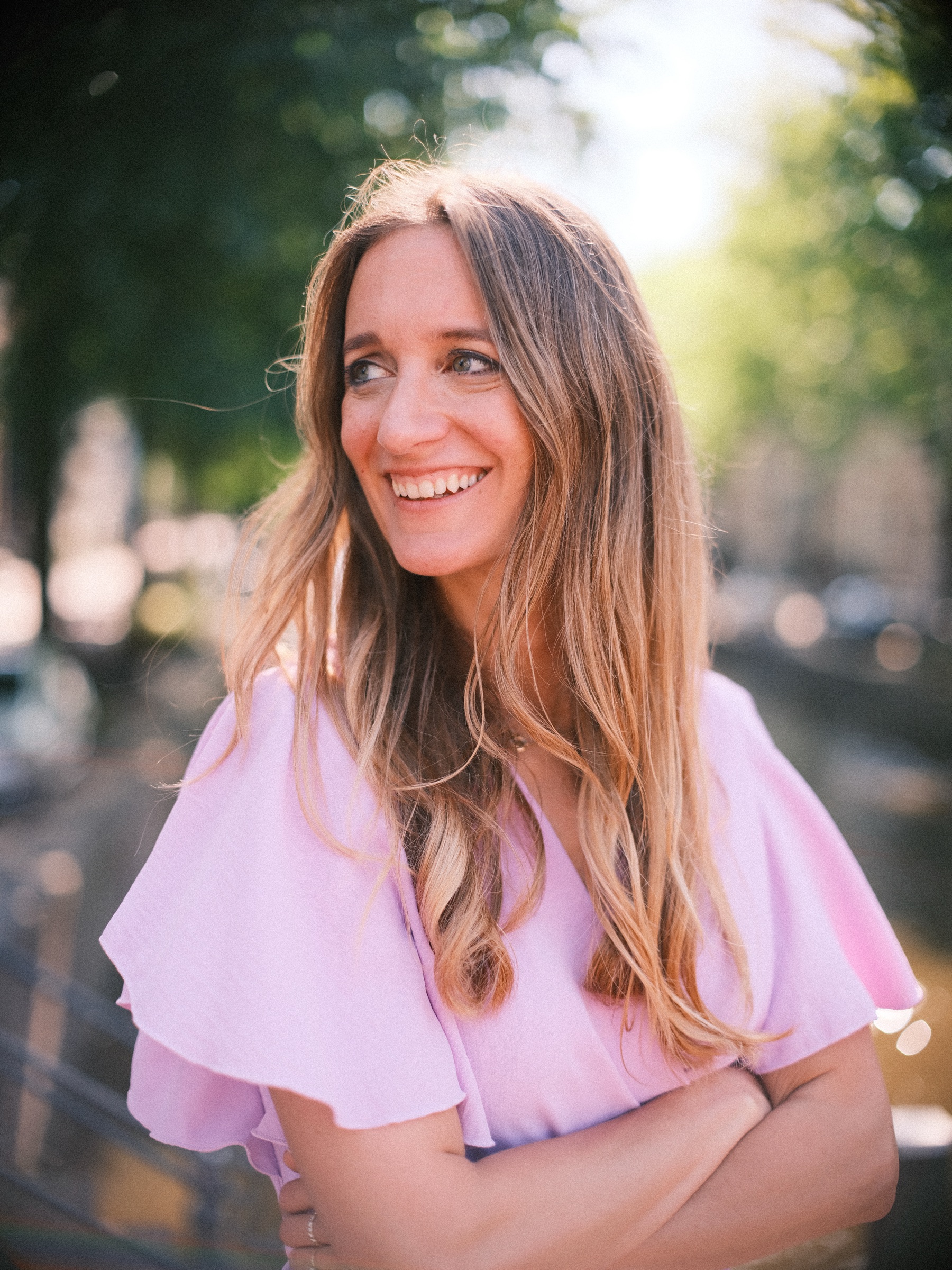 Close portrait of the bride-to-be smiling to one side, arms crossed over her lilac dress, her long hair lit by the summer sun