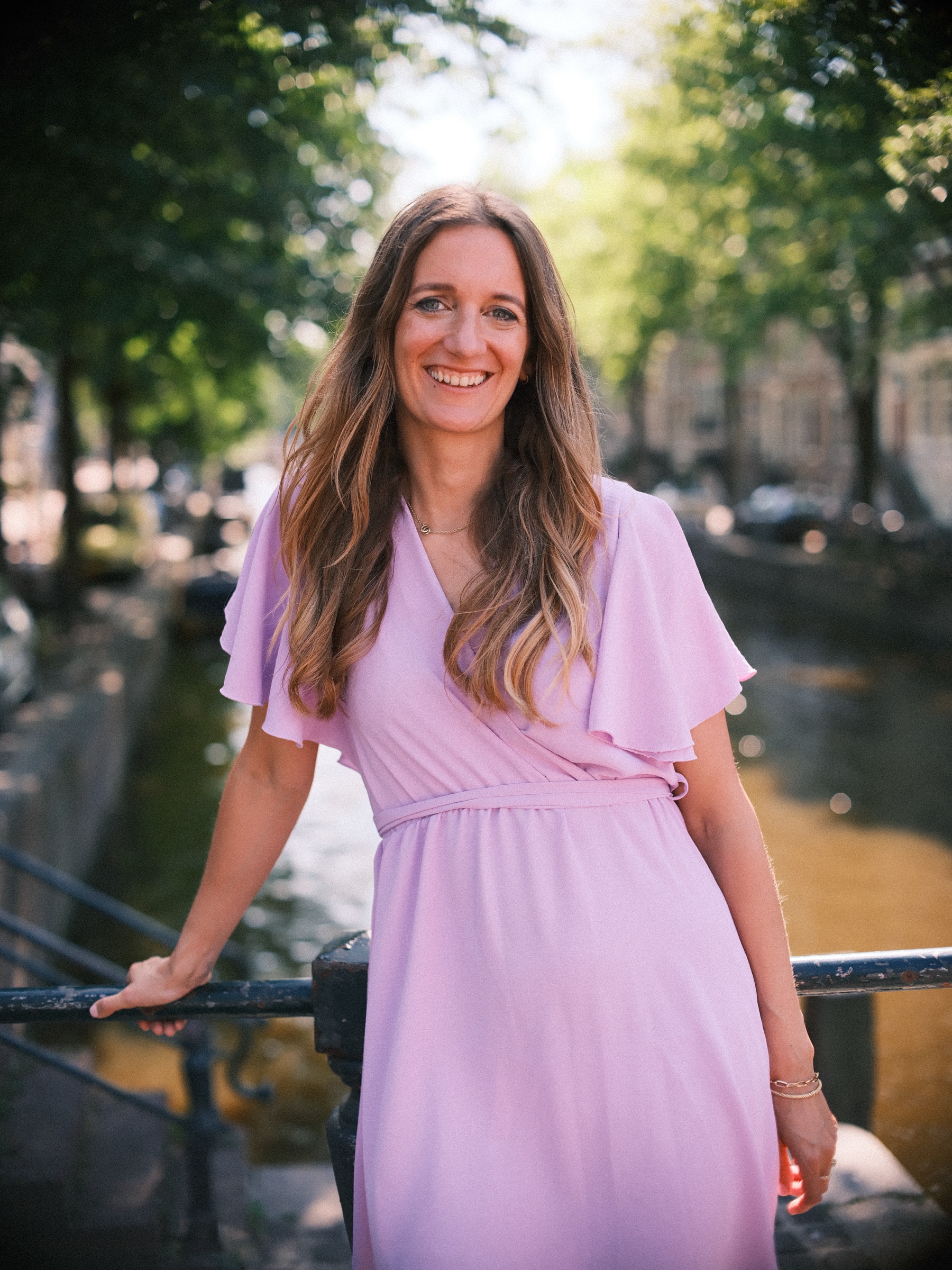 Bride-to-be in a lilac dress with fluttering sleeves leaning on a canal bridge railing, trees and canal houses behind her
