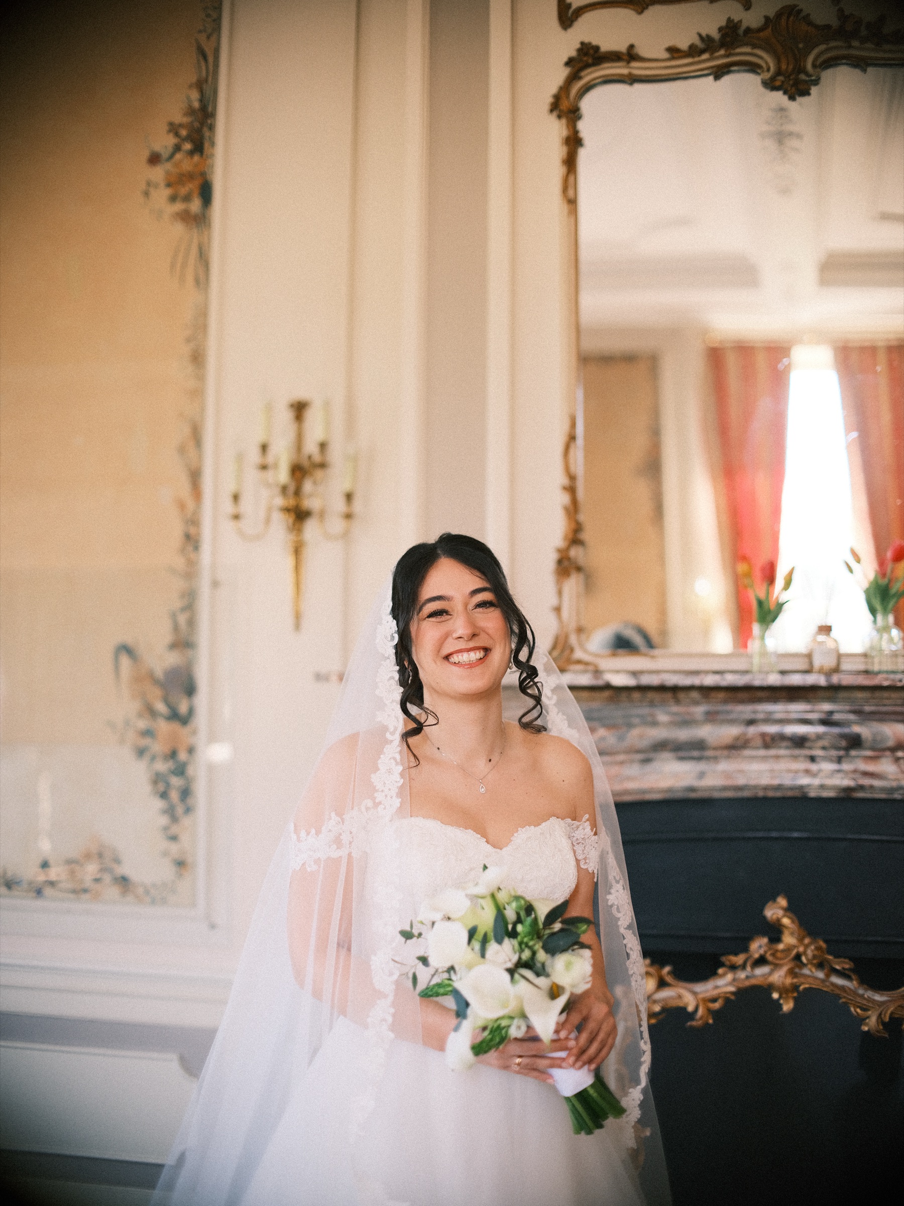 Bruid in een jurk met blote schouders en een lange sluier met kant lacht bij een marmeren schouw, met witte calla's in haar handen