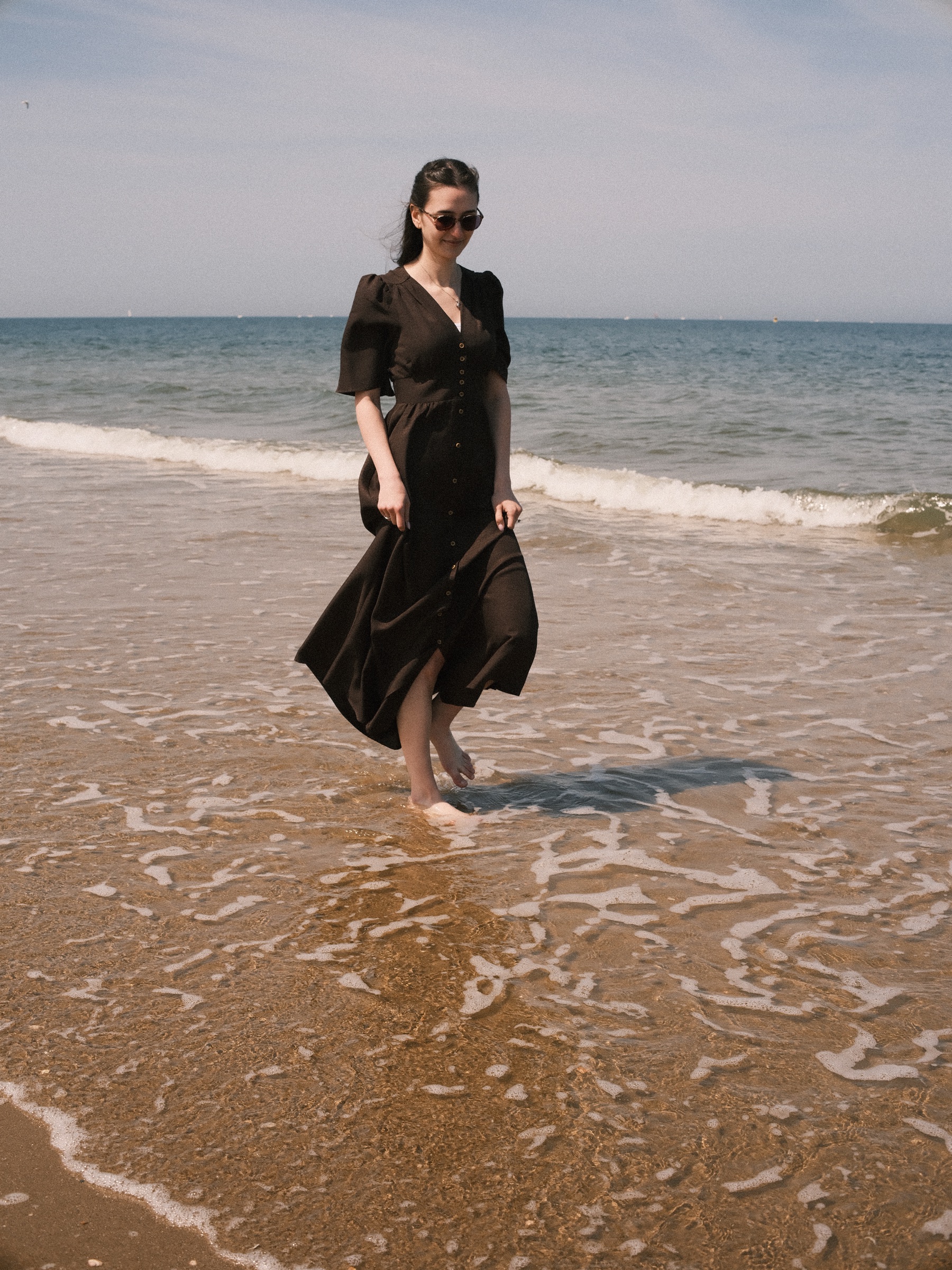 Woman in a long dark brown dress walking barefoot through the shallow surf, the water curling around her feet