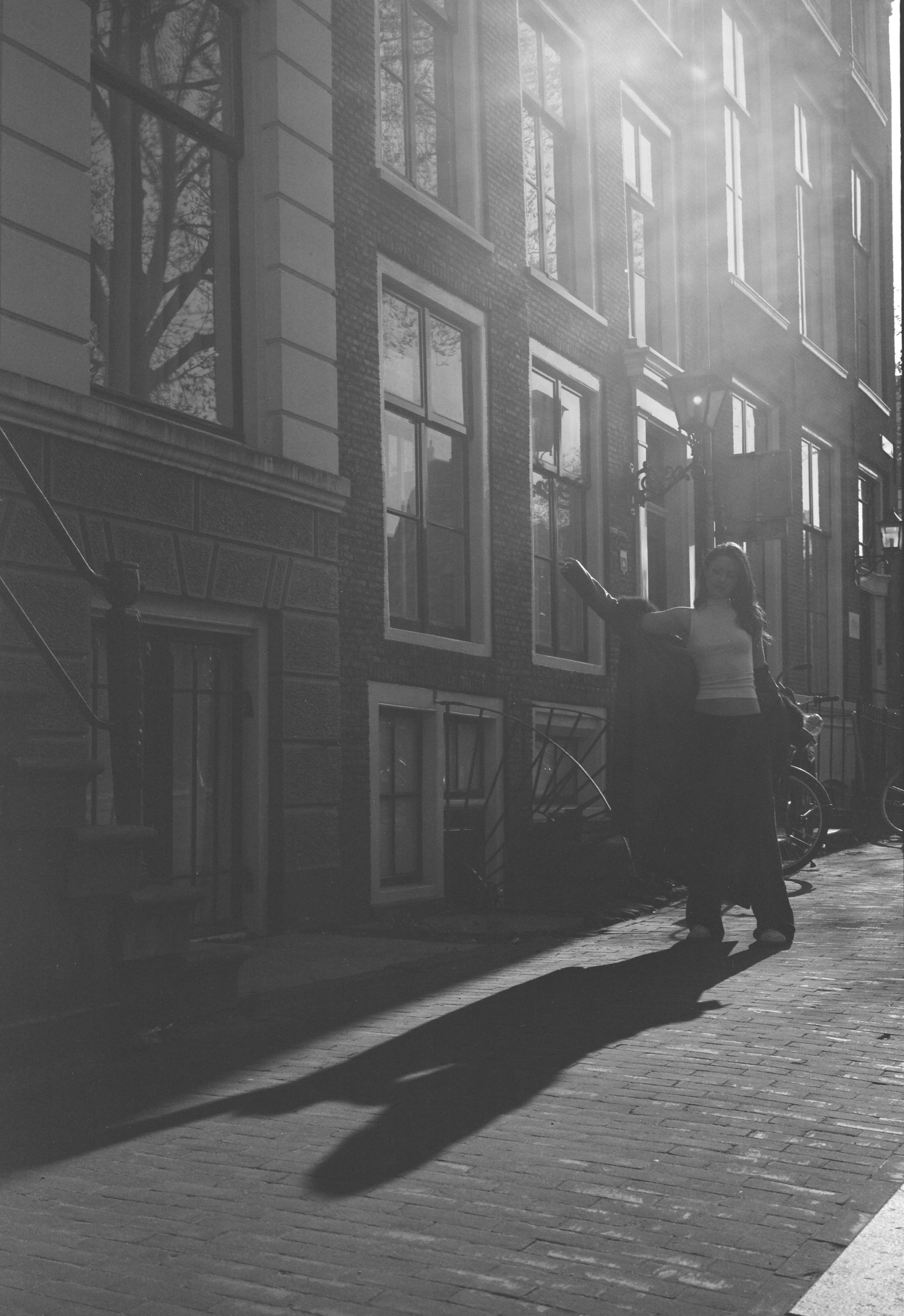Zonnige gevel van een grachtenpand met hoge ramen, een vrouw bij de stoep en haar lange schaduw op de stenen, op Fomapan 100