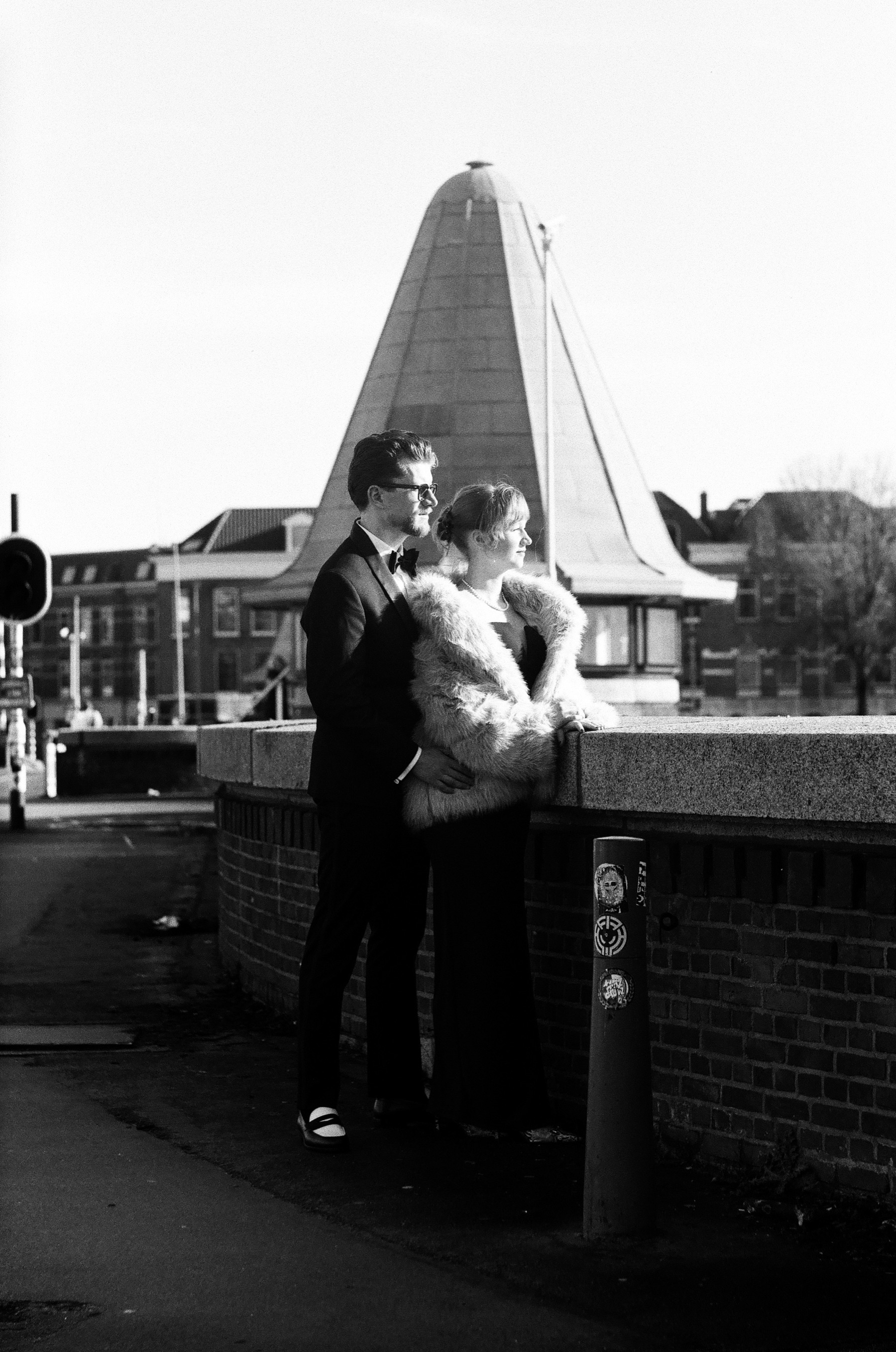 Couple in a tuxedo and a pale fur coat standing at a brick parapet in Rotterdam, faces turned to the sun, a building with a tall pointed roof behind them