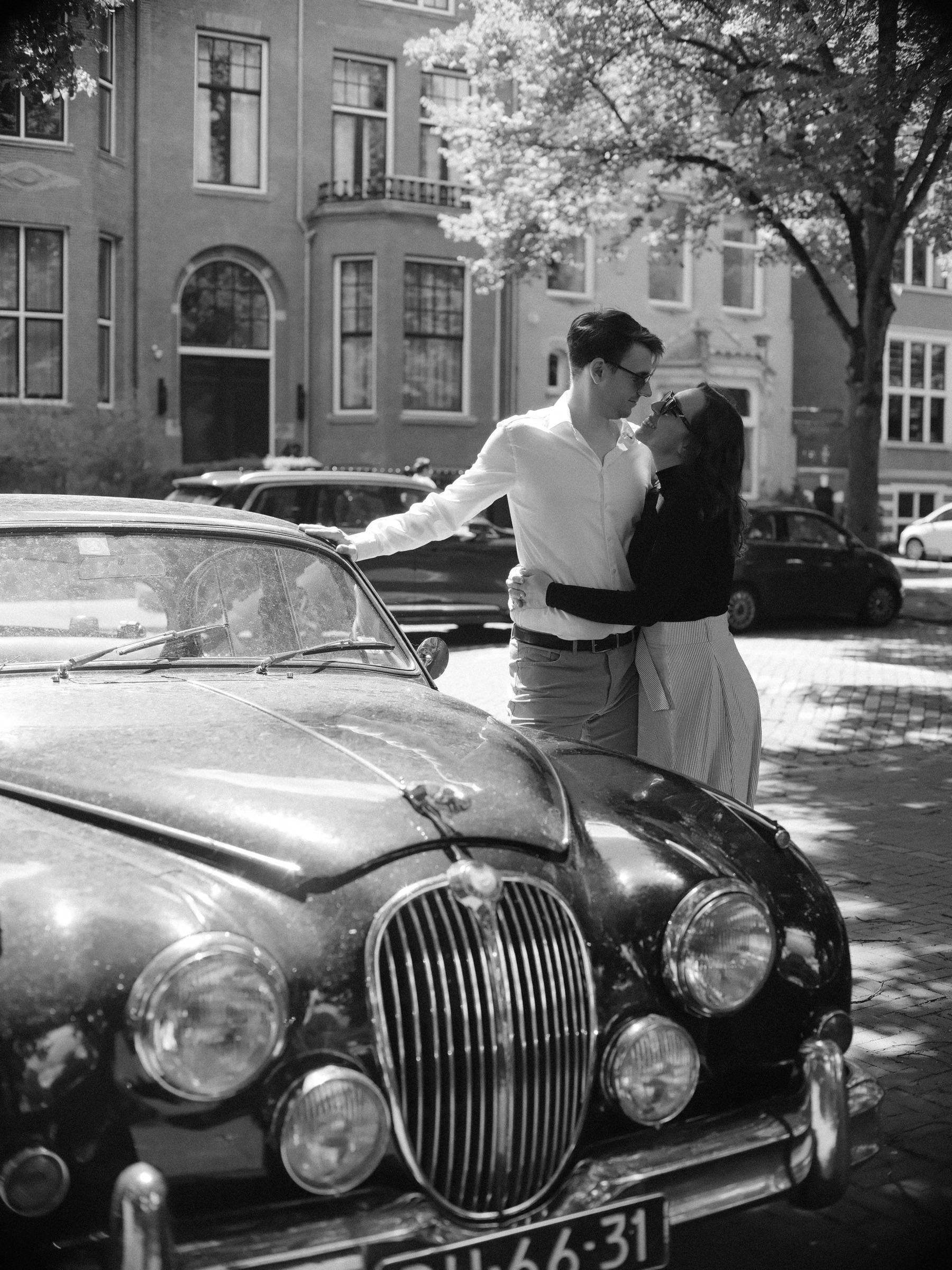 Classic Jaguar with its chrome grille in the foreground, the couple embracing beside its roof under leafy trees, in black and white