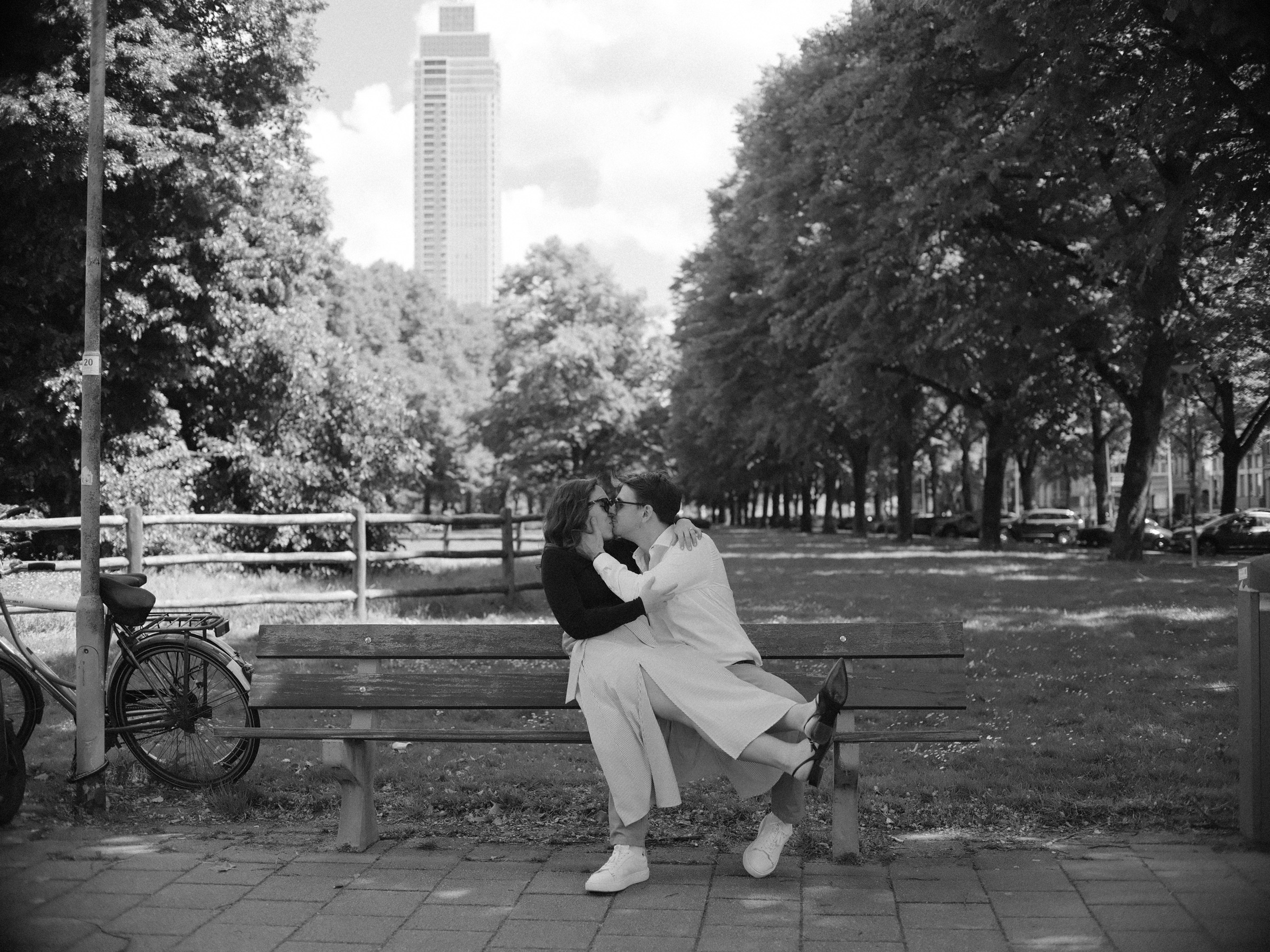 Black and white frame of a couple kissing on a wooden park bench, a bike and a tall tower behind, the end of a photo walk in Rotterdam