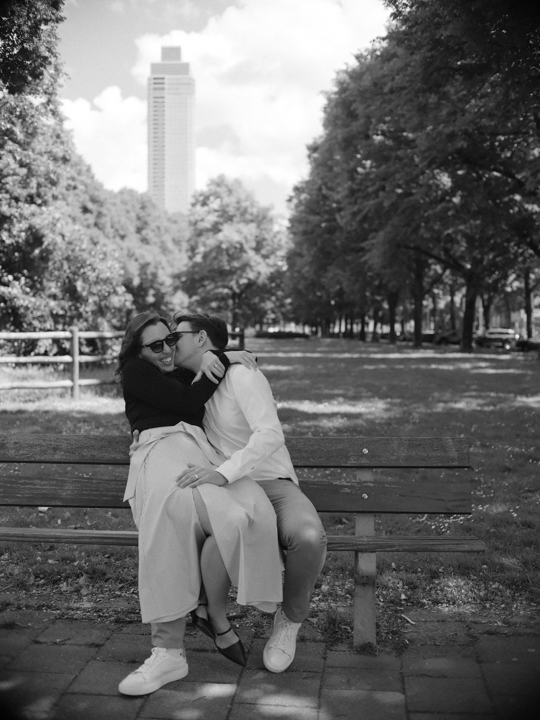 On a wooden park bench she sits on his lap and kisses his cheek, a long row of trees and a tall tower behind them, in black and white