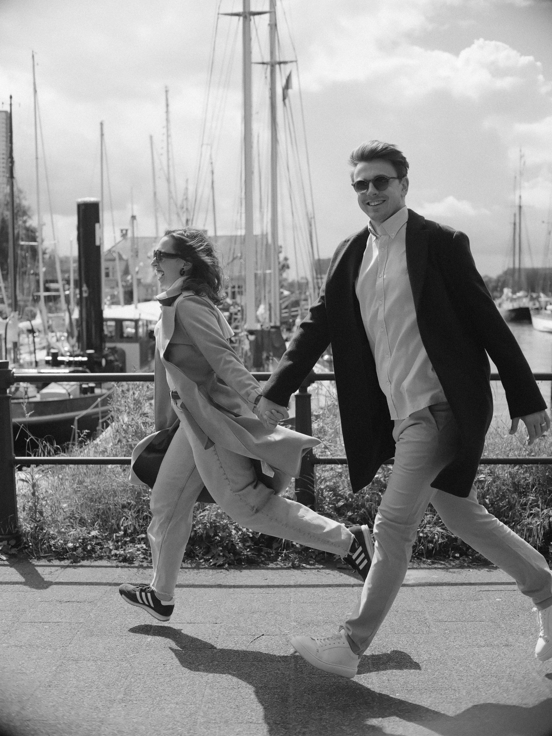 Black and white frame of the couple running hand in hand along the harbor railing, long coats flying and sharp shadows on the stones
