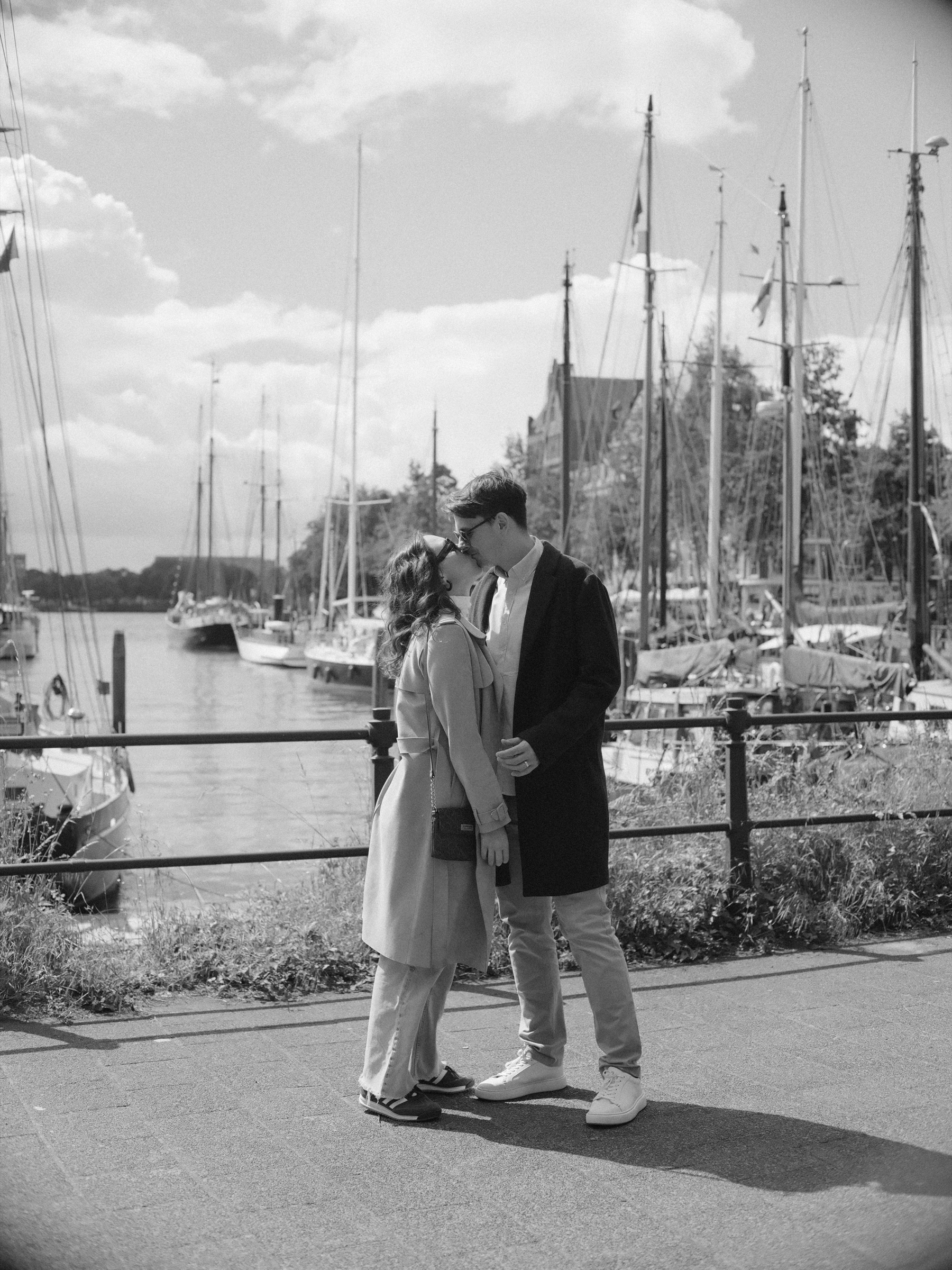 Couple kissing by the railing of the Veerhaven in Rotterdam during a digital photoshoot in black and white, sailing ship masts behind them
