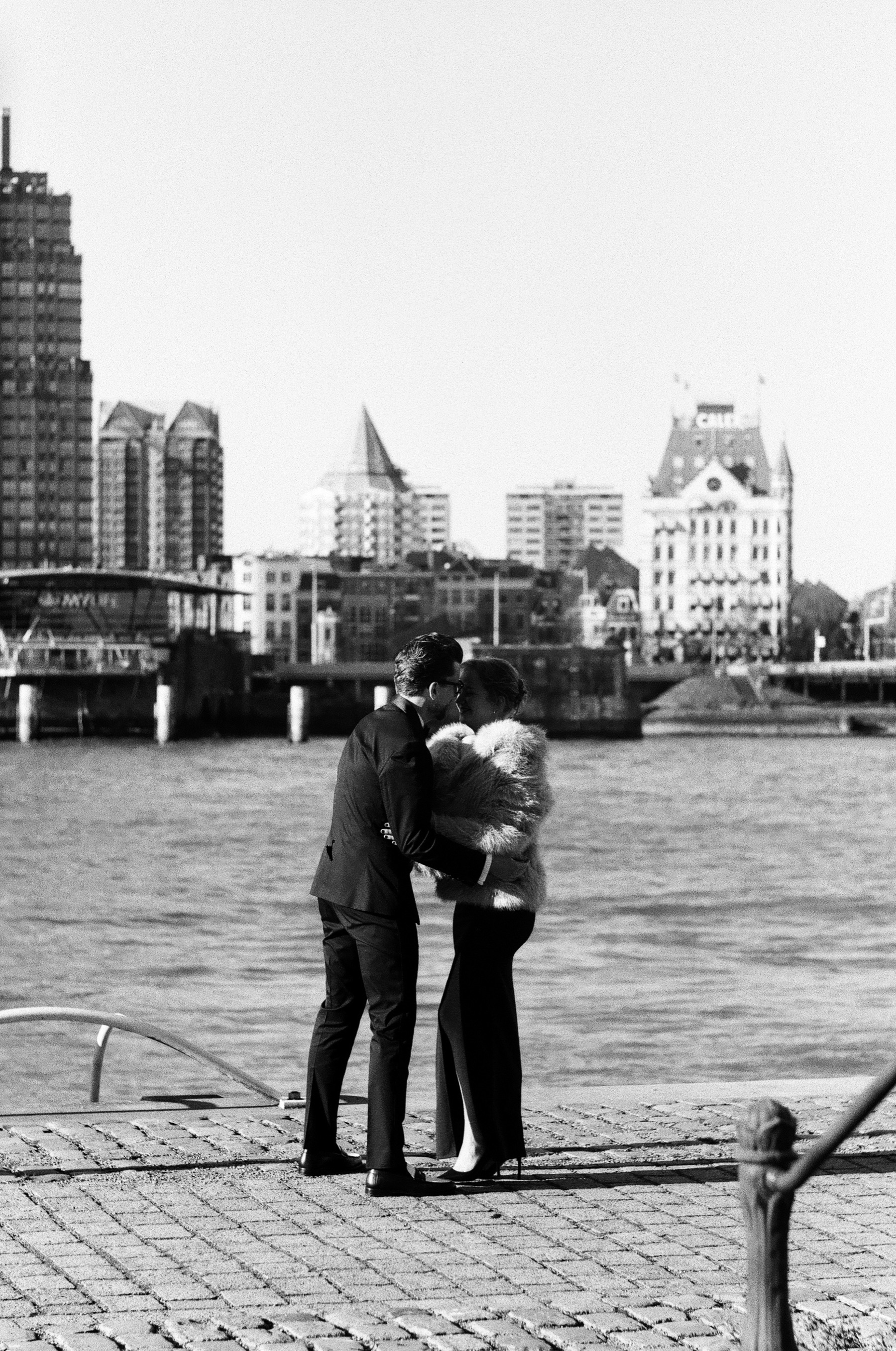 Couple kissing on the cobbled quay of the Maas in Rotterdam, Het Witte Huis and tall towers across the water, a black and white film photoshoot