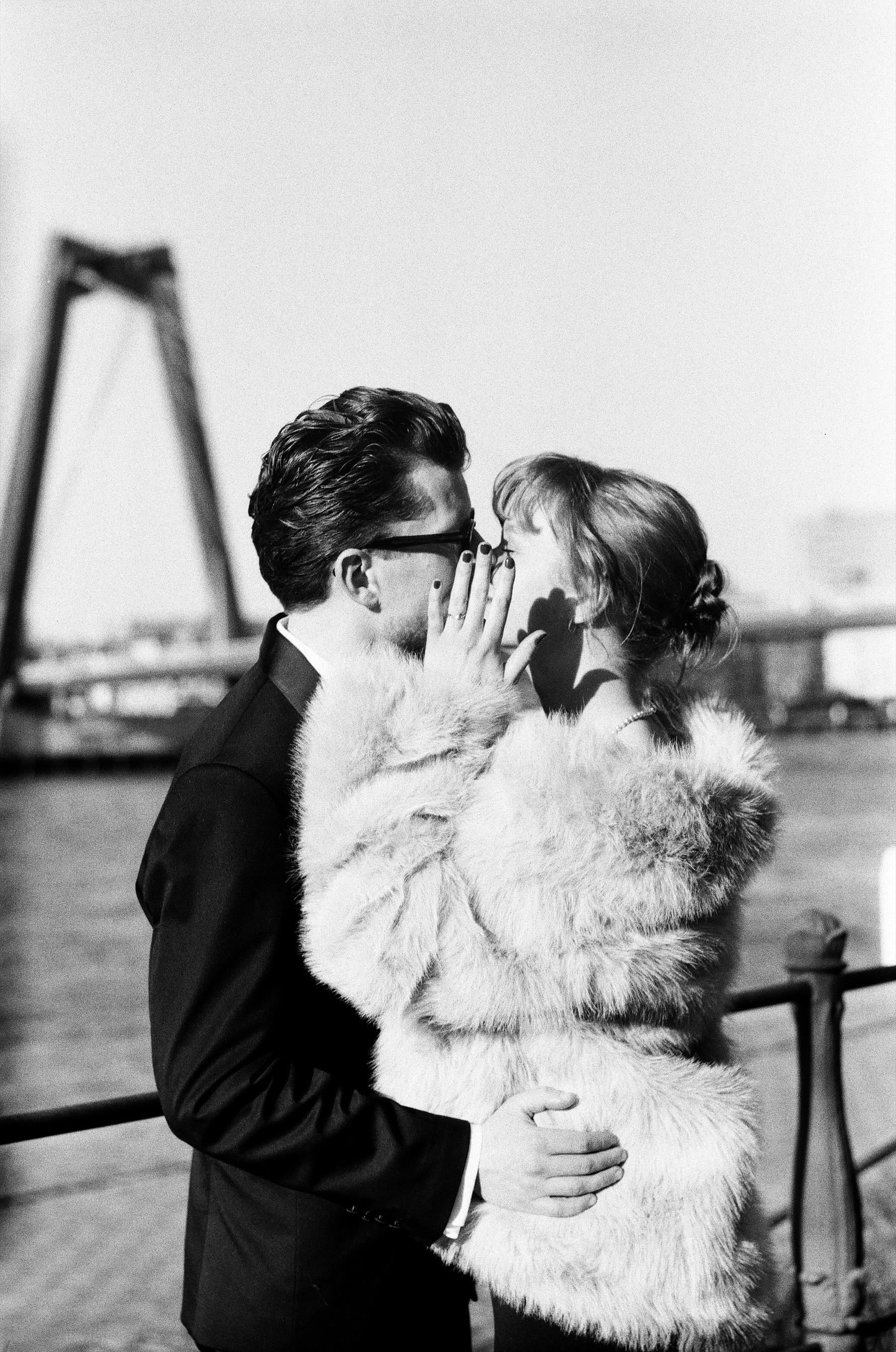Close kiss on black and white film, her hand on his cheek and her fur coat filling the frame, the pylon of the Willemsbrug behind