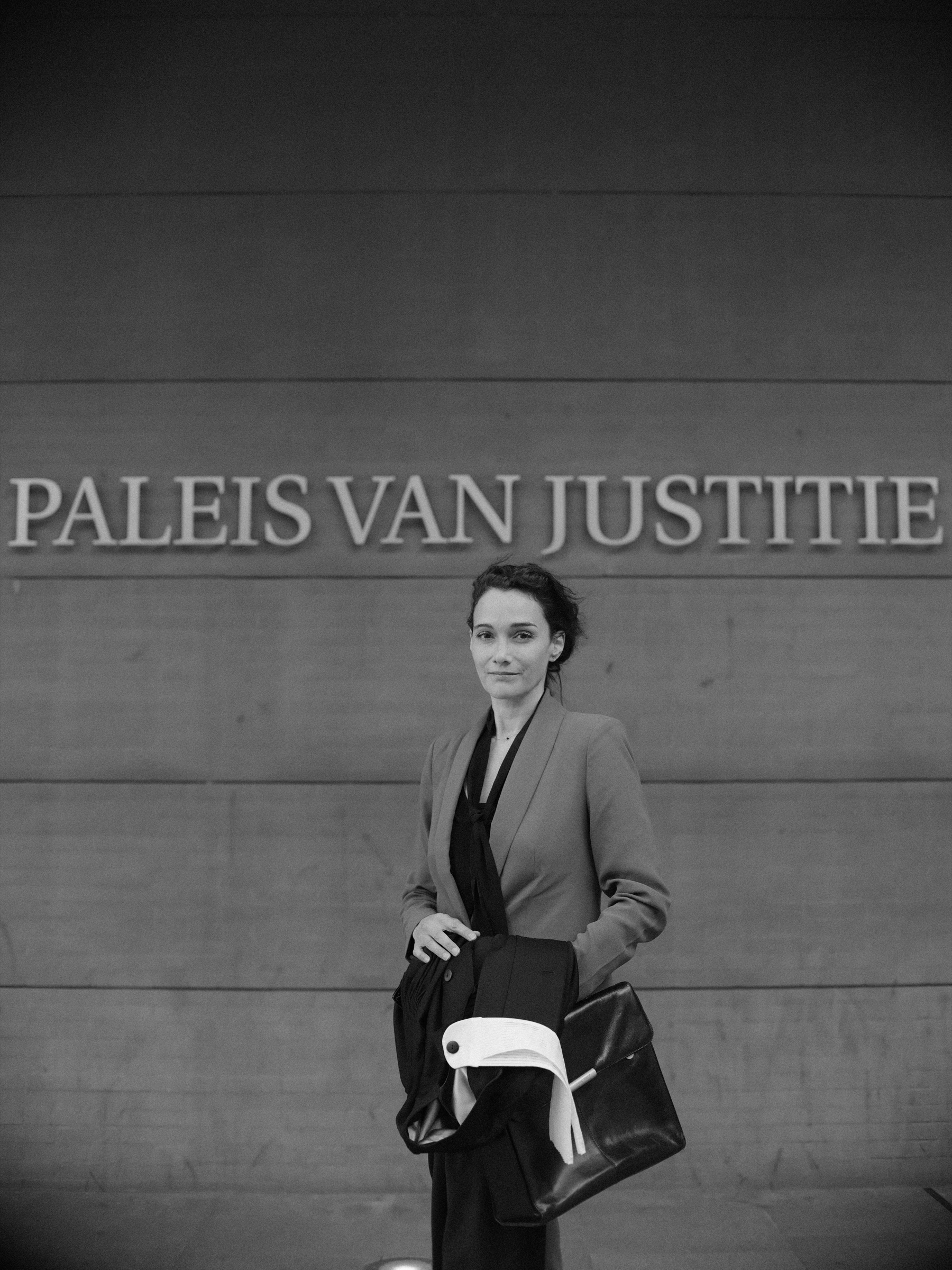 Zwart-witportret van een vrouw onder de letters Paleis van Justitie in Den Haag, met een zwarte toga met witte bef over haar arm