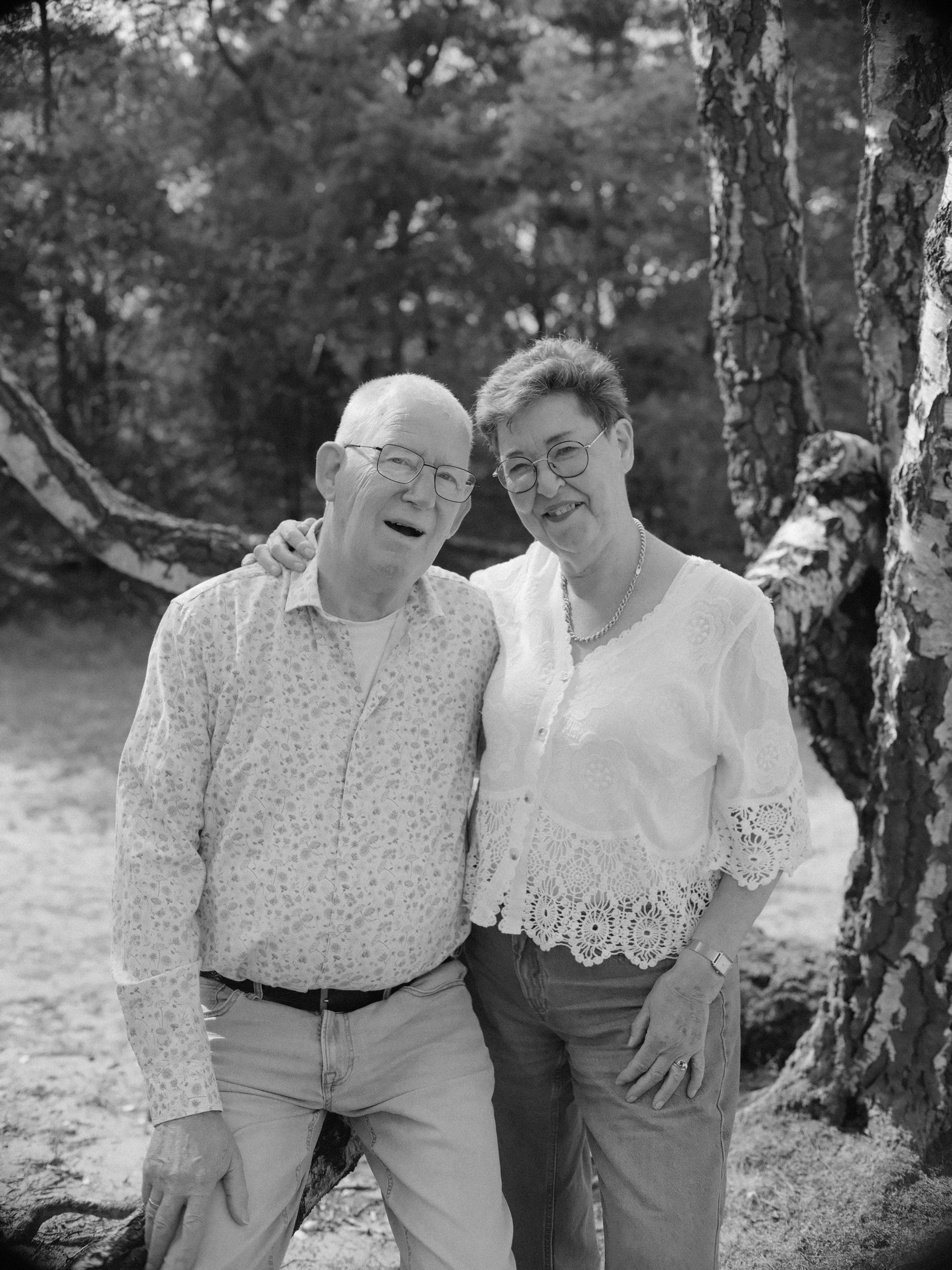 Black and white portrait of a grandfather laughing with his arm around his wife in a lace blouse, birch trunks behind them