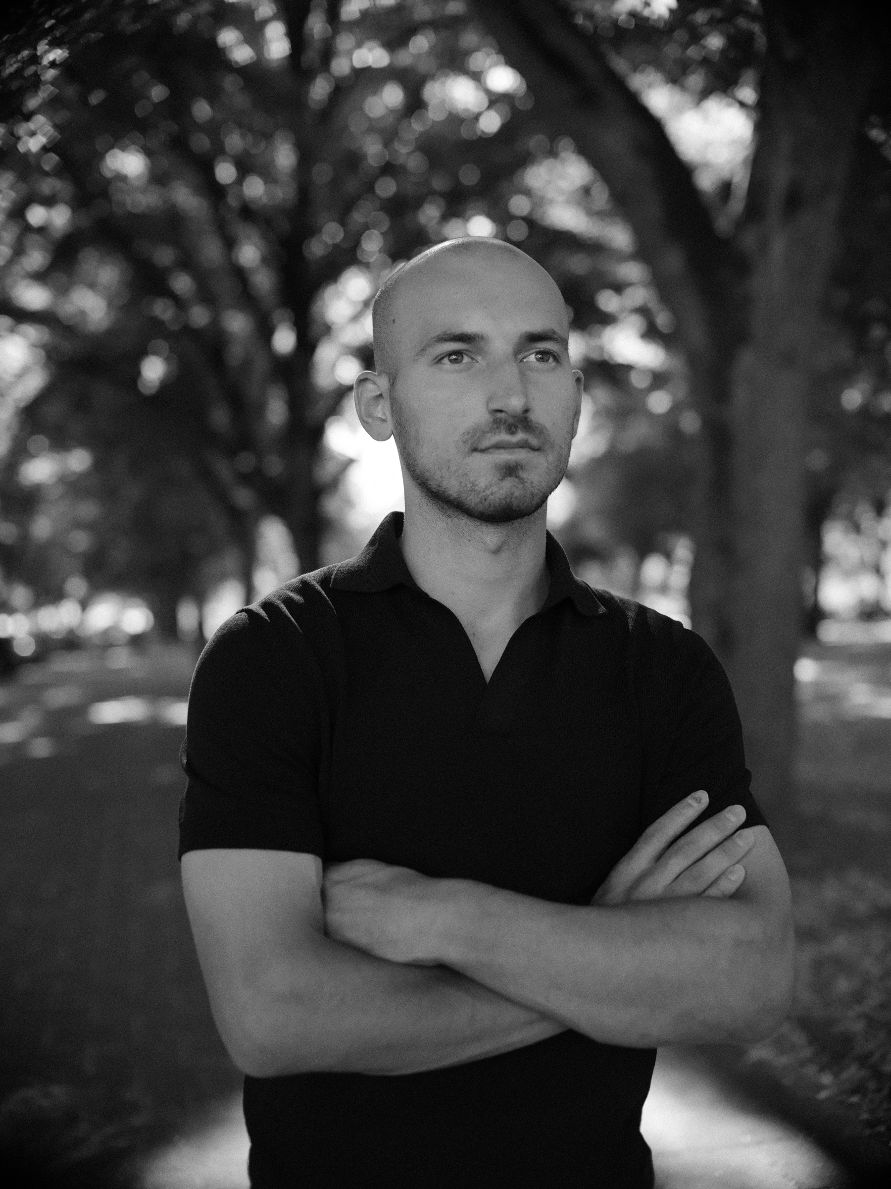 Zwart-witportret met de armen over elkaar voor een lange laan met bomen, terwijl zacht licht door de bladeren valt